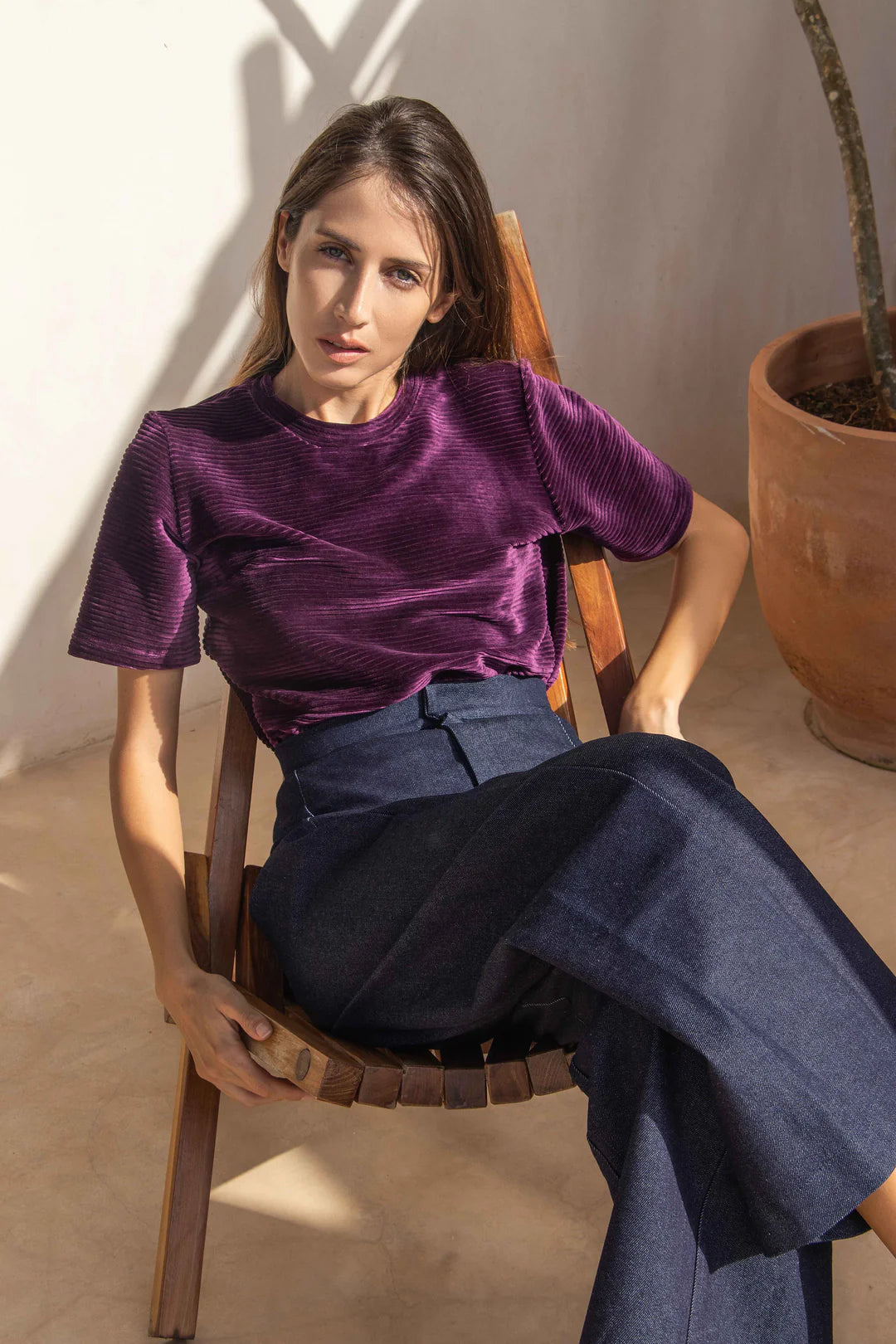 A woman wearing the Vienna Pants by MAS in Jeans, with a wide leg, high waist, side seams that sit slightly to the front, and two back pockets, with a purple top. She is sitting in a wooden chair.