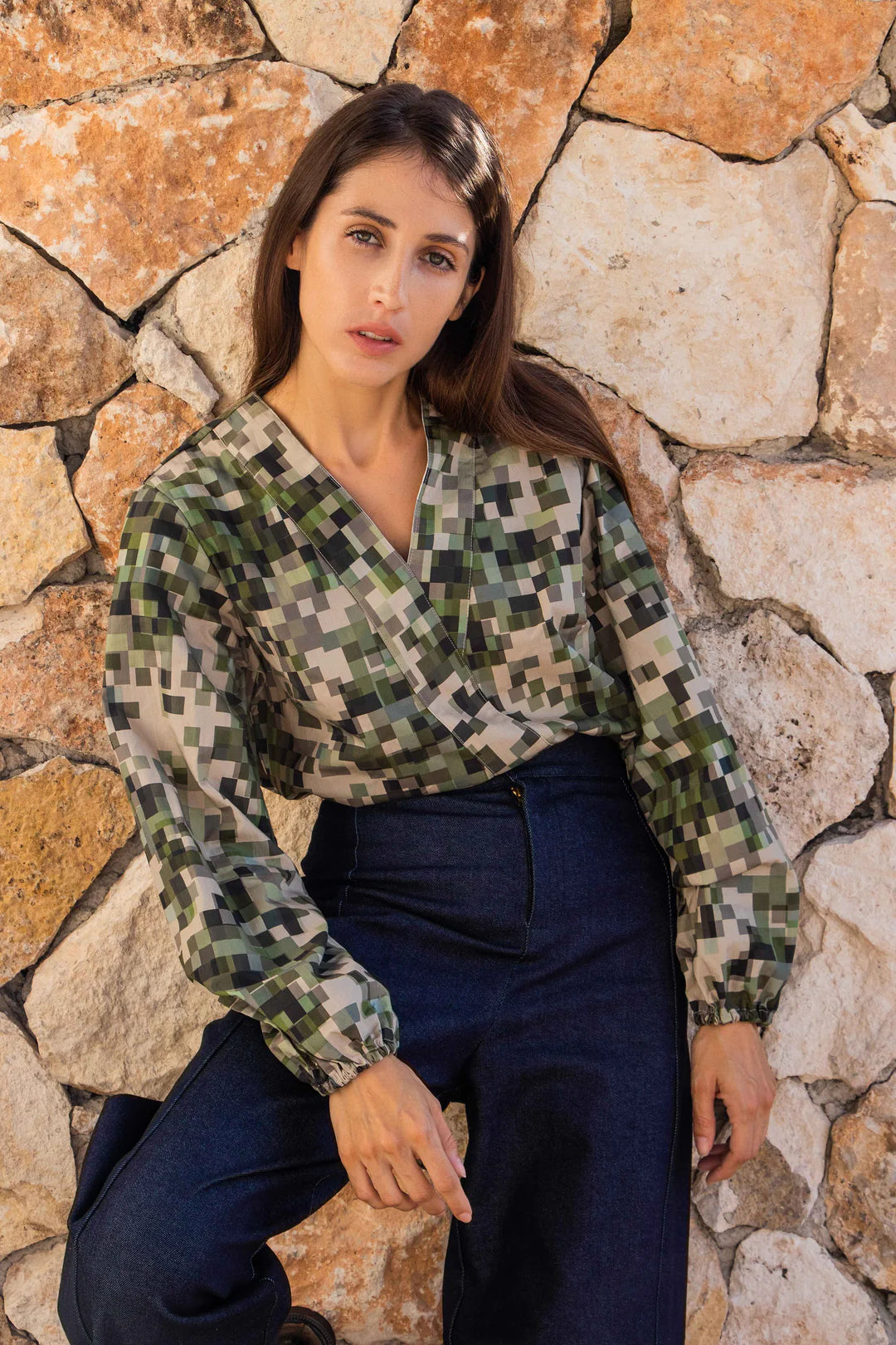 A woman wearing the Vienna Pants by MAS in Jeans, high waisted, wide-legged pants with a zipper and tab. She is wearing them with a green top and standing in front of a stone wall.
