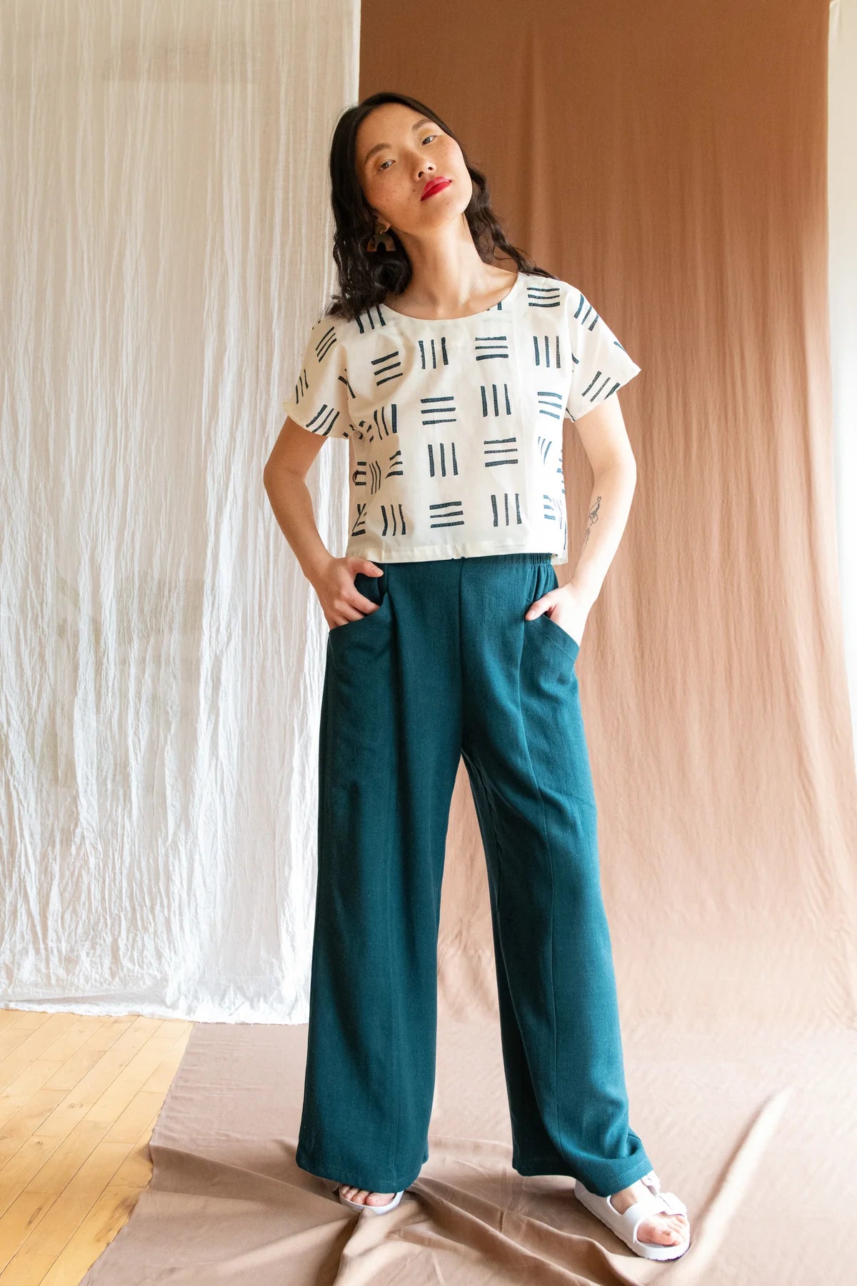 A woman wearing the Padua Pants by Kazak in Forest with a high waist, flat waistband at the front and elastic at the bag, wide legs, and large slanted pockets. She is wearing them with a patterned top and standing in front of a brown curtain.