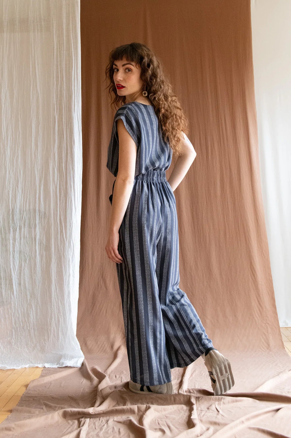 Side/back view of 3/4 view of A woman wearing the Padua Pants by Kazak in Charcoal Dots, with a high waist, flat waistband at the front and elastic at the bag, wide legs, and large slanted pockets. She is wearing them with the matching Sandrigo Top and standing in front of a brown curtain.