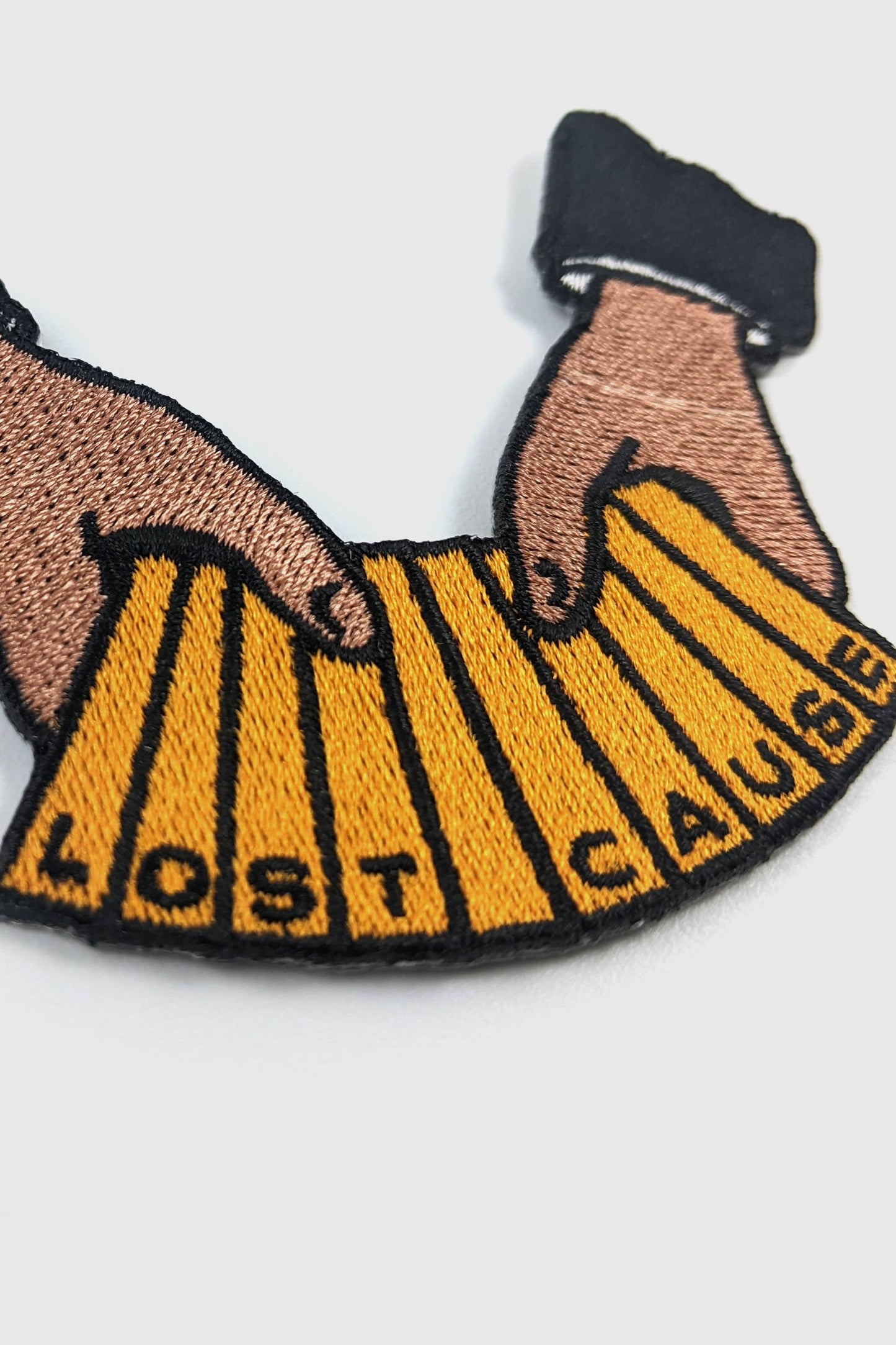 An embroidered patch by Badaboöm Studio depicting two hands holding a yellow accordion that reads 'LOST CAUSE' in black letters, with dark blue cuffs, on a light grey background.