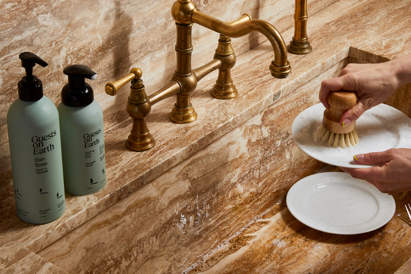 Person washing dishes with a brush using Guests on Earth dish soap, next to the bottle of soap on a marble countertop.