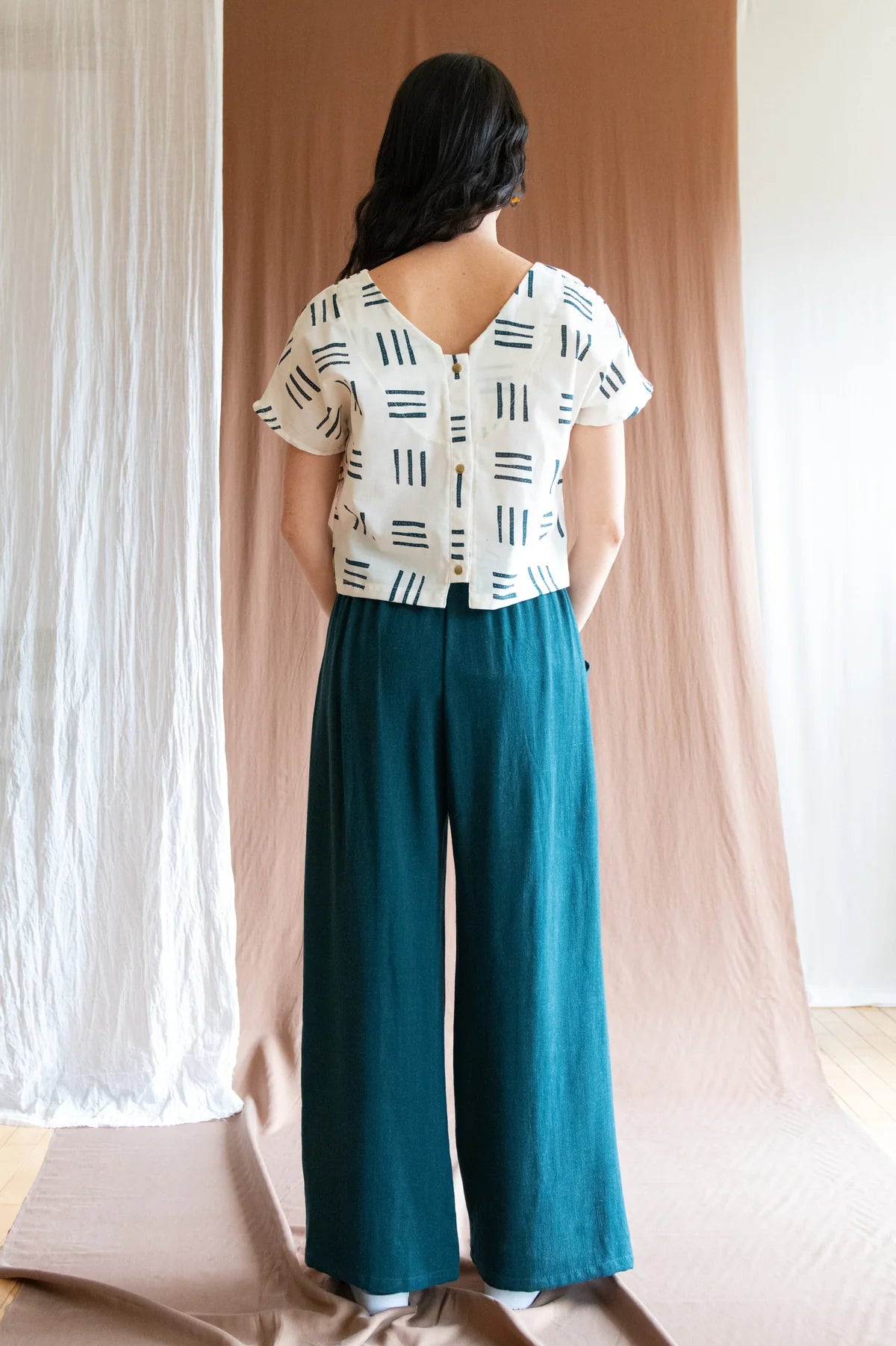 Back view of a womanwearing the Padua Pants by Kazak in Forest, with a high waist, flat waistband at the front and elastic at the bag, wide legs, and large slanted pockets. She is wearing them with a printed top and standing in front of a brown curtain.