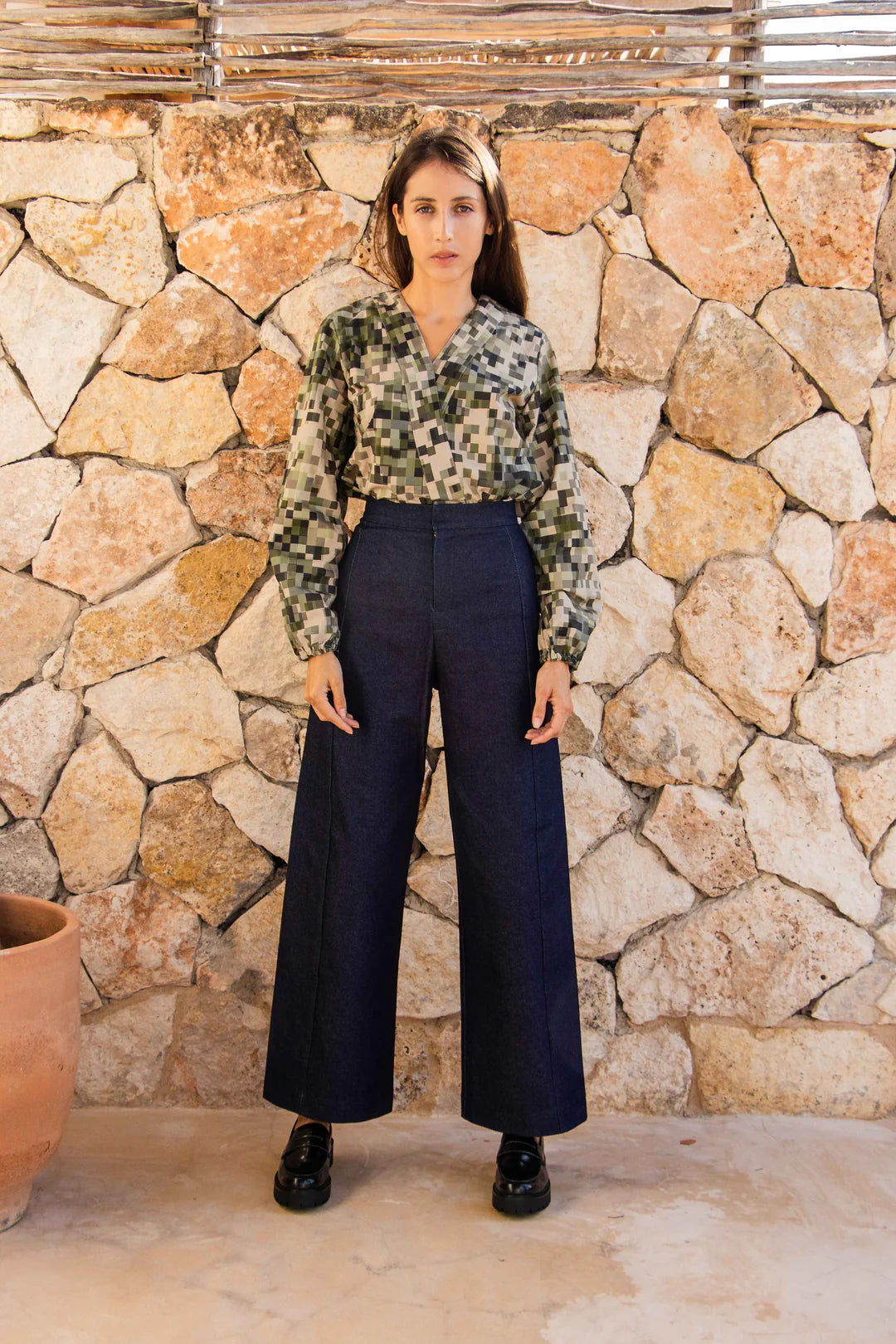 A woman wearing the Vienna Pants in Jeans by MAS, high waisted pants with wide legs and a zipper and tab closure. She is wearing them with a green top and standing in front of a stone wall.