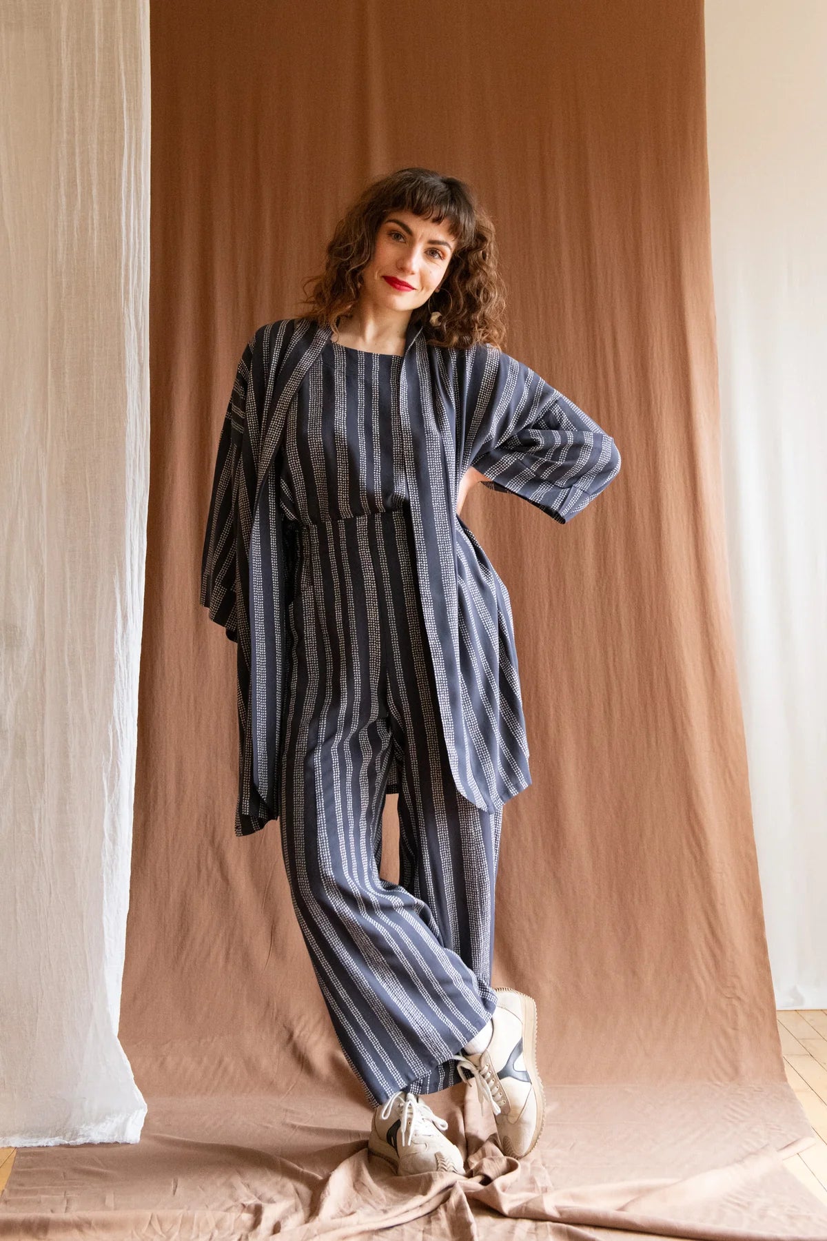 A woman wearing the Padua Pants by Kazak in Charcoal Dots, with a high waist, flat waistband at the front and elastic at the bag, wide legs, and large slanted pockets. She is wearing them with the matching Sandrigo top and Mirano Jacket and standing in front of a white curtain.