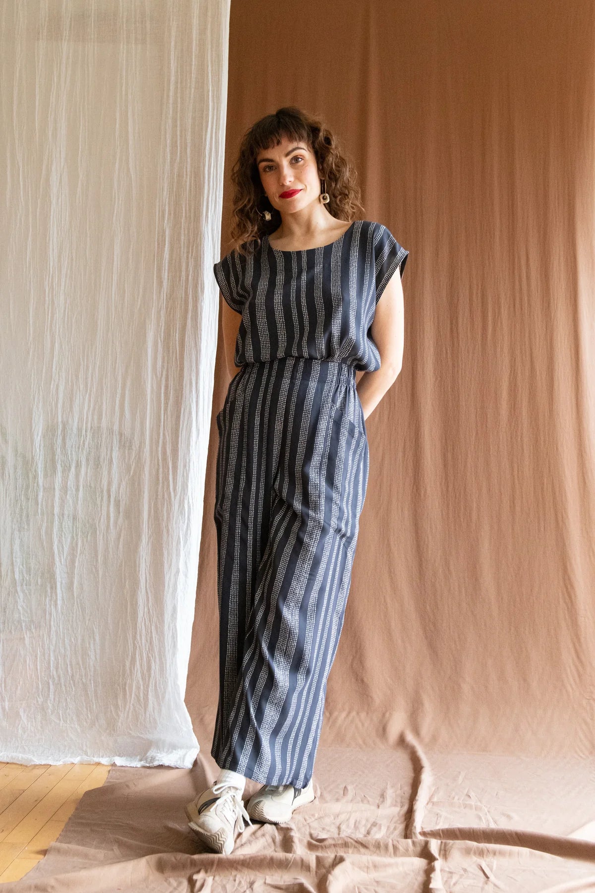 A woman wearing the Padua Pants by Kazak in Charcoal Dots, with a high waist, flat waistband at the front and elastic at the bag, wide legs, and large slanted pockets. She is wearing them with the matching Sandrigo Top and standing in front of a brown curtain.