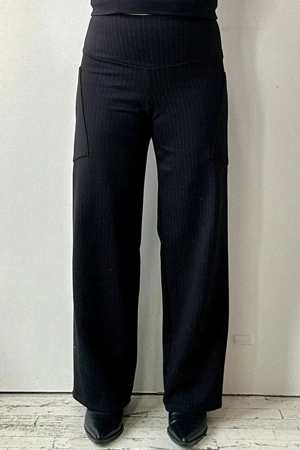 Waist-down view of a woman wearing the Susan Pants by Kollontai in Black pinstripe, with wide legs, a wide pull-on waistband, and angled side patch pockets. She is standing in front of a white wall.