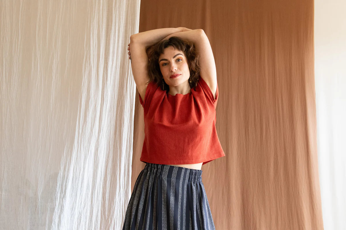 3/4 view of a woman wearing the Brendola Top by Kazak in Rust, a reversible top with a v-neck and snap front on one side and a round neck on the other side. It has short extended sleeves and a relaxed, cropped fit. She is wearing it with a patterned skirt and standing in front of a brown curtain.