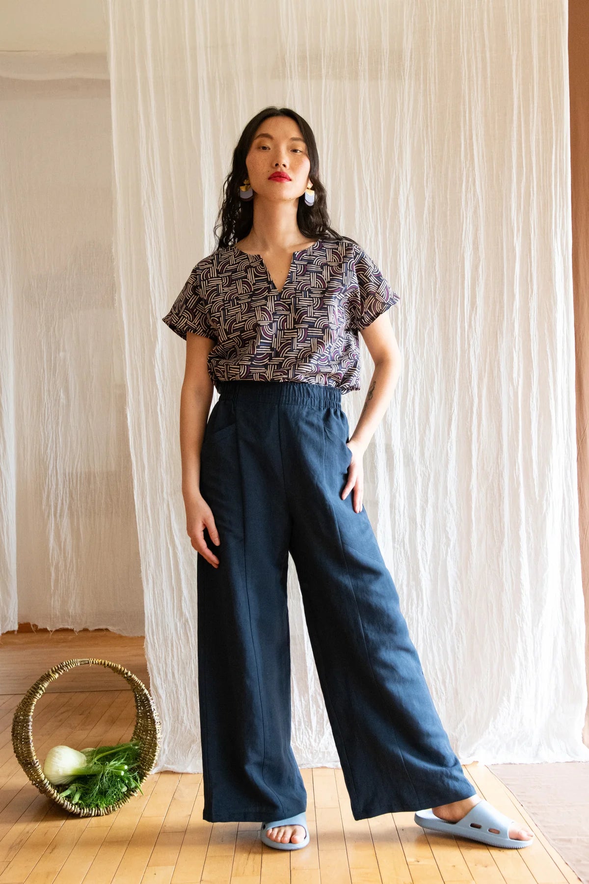 A woman wearing the Padua Pants by Kazak in Indigo, with a high waist, flat waistband at the front and elastic at the bag, wide legs, and large slanted pockets. She is wearing them with the Lucca Top in Night Basketwork and standing in front of a white curtain.