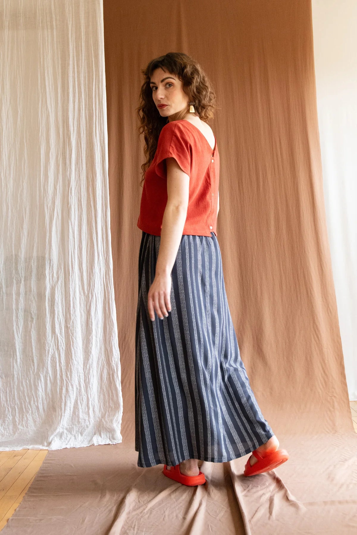 Side/back view of a woman wearing the Brendola Top by Kazak in Rust, a reversible top with a v-neck and snap front on one side and a round neck on the other side. It has short extended sleeves and a relaxed, cropped fit. She is wearing it with the Verona Skirt in Charcoal Dots and standing in front of a brown curtain.