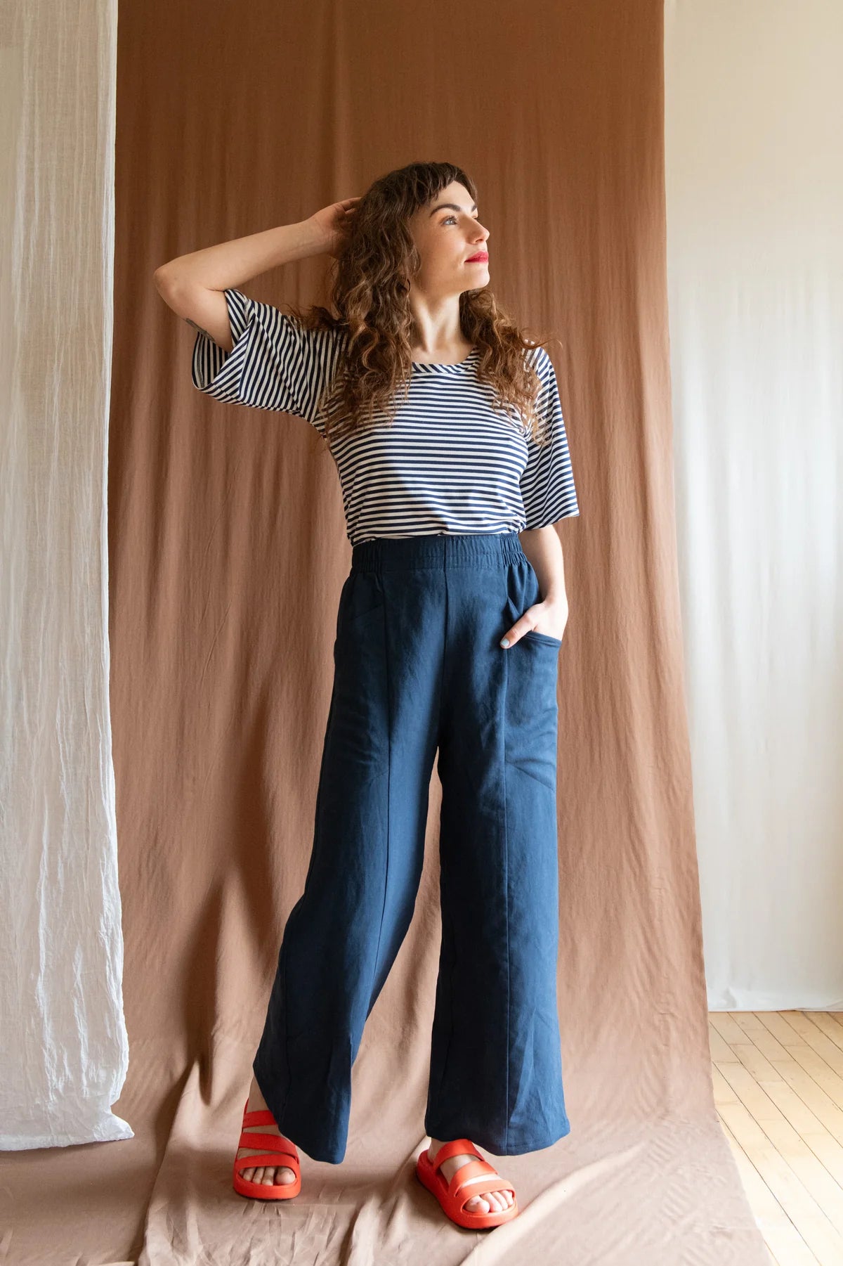 A woman wearing the Padua Pants by Kazak in Indigo, with a high waist, flat waistband at the front and elastic at the back, wide legs, and large slanted pockets. She is wearing them with the striped Montebelluna top and standing in front of a brown curtain.