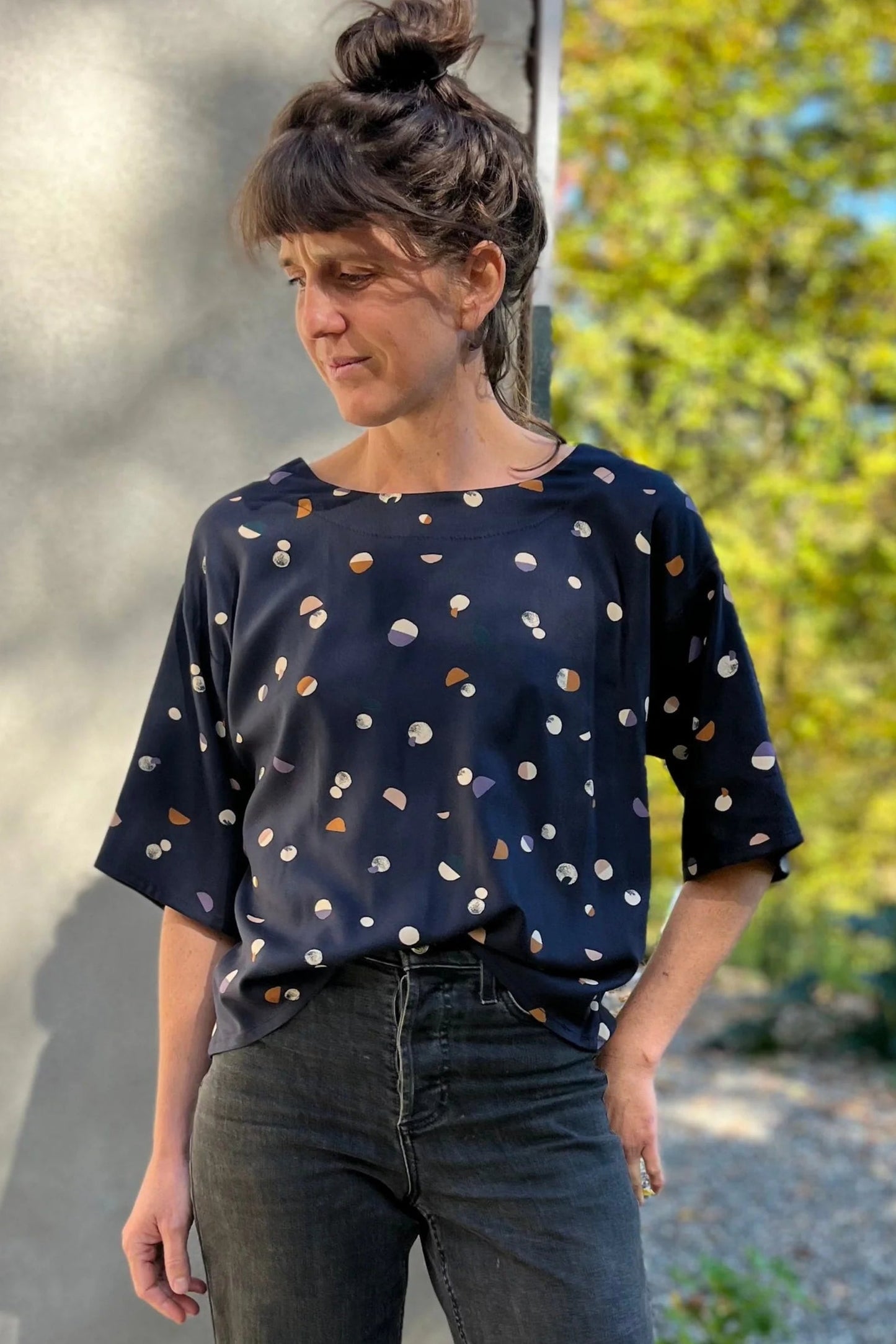 A woman wearing the Abelia Top by Kazak in Midnight Moons, a boxy shirt with a wide neck and 3/4 kimono sleeves. She is wearing it with grey jeans and standing outside against a grey wall.