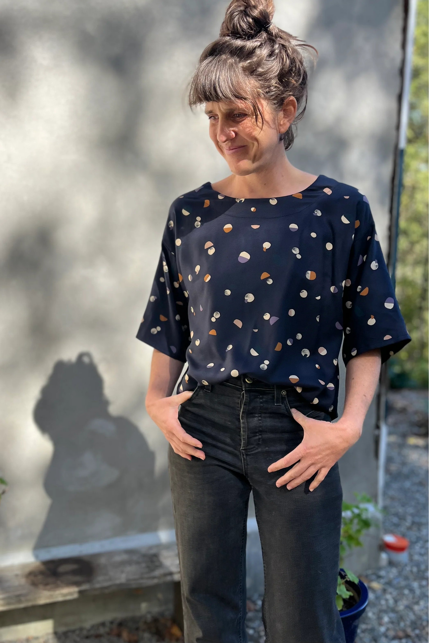 A woman wearing the Abelia Top by Kazak in Midnight Moons, a boxy shirt with a wide neck and 3/4 kimono sleeves. She is wearing it with grey jeans and standing outside against a grey wall.