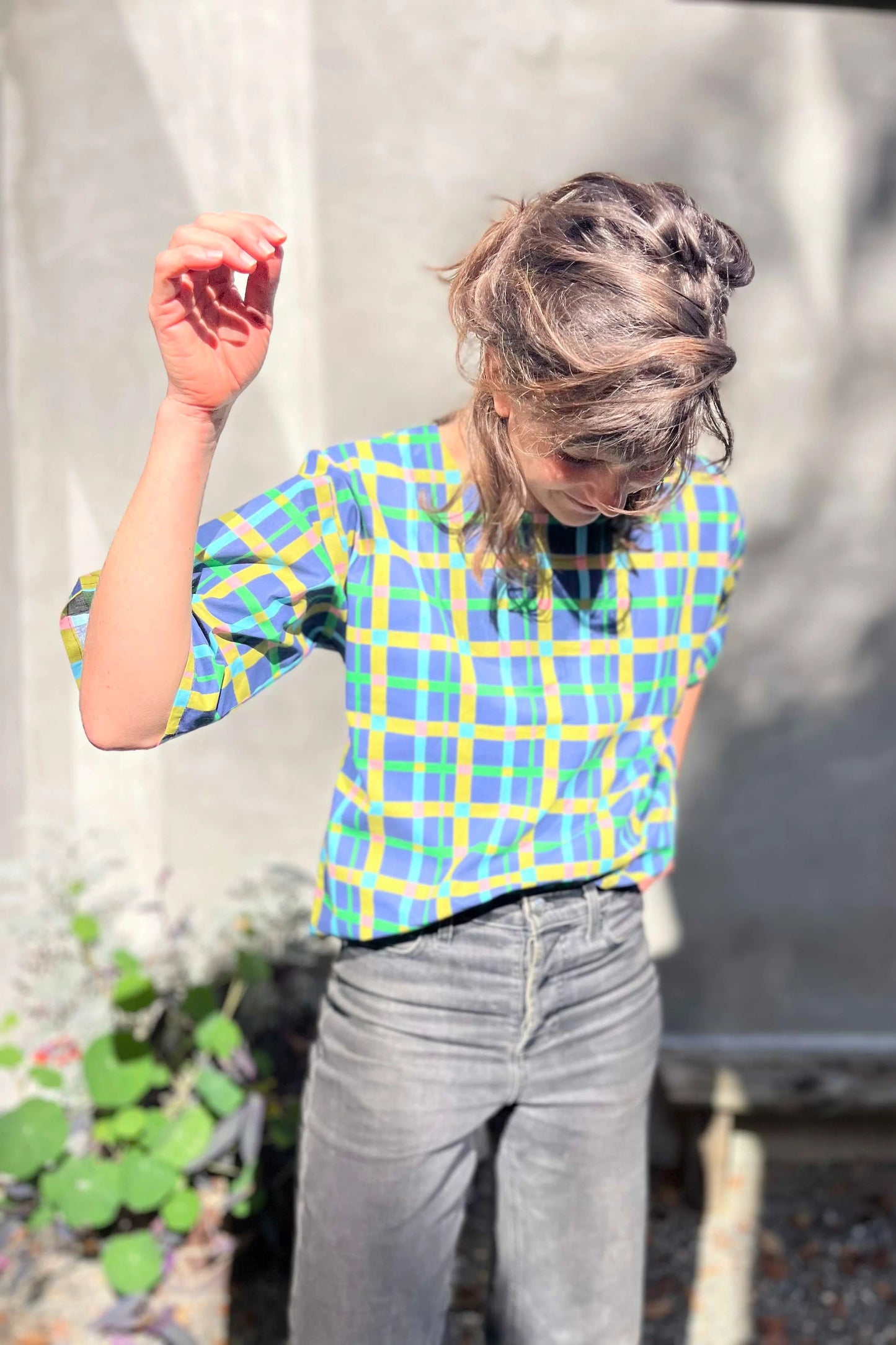A woman wearing the Abelia Top by Kazak in Blue checks, a boxy shirt with a wide neck and 3/4 kimono sleeves. She is wearing it with grey jeans and standing outside against a grey wall.