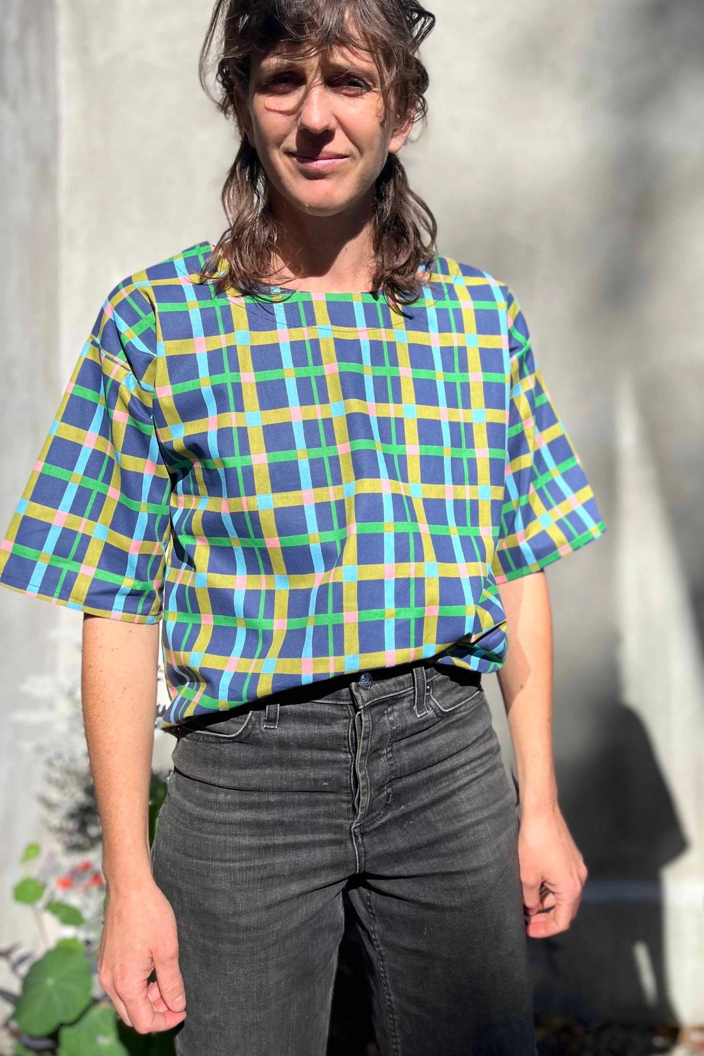 A woman wearing the Abelia Top by Kazak in Blue checks, a boxy shirt with a wide neck and 3/4 kimono sleeves. She is wearing it with grey jeans and standing outside against a grey wall.
