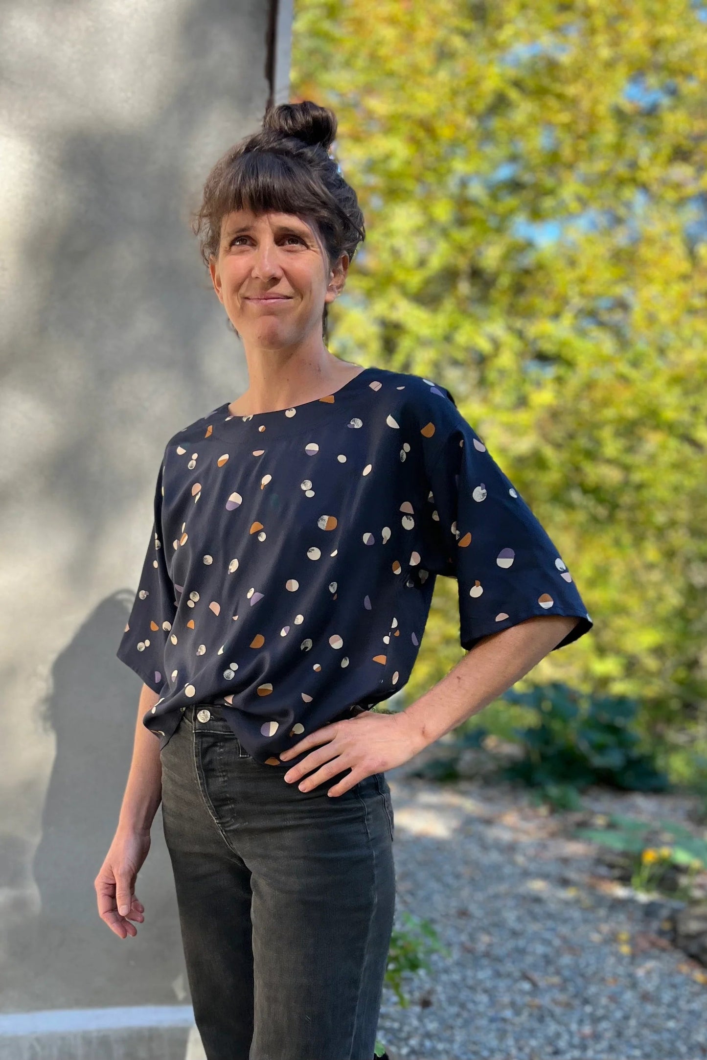 A woman wearing the Abelia Top by Kazak in Midnight Moons,a boxy shirt with a wide neck and 3/4 kimono sleeves. She is wearing it with grey jeans and standing outside against a grey wall.