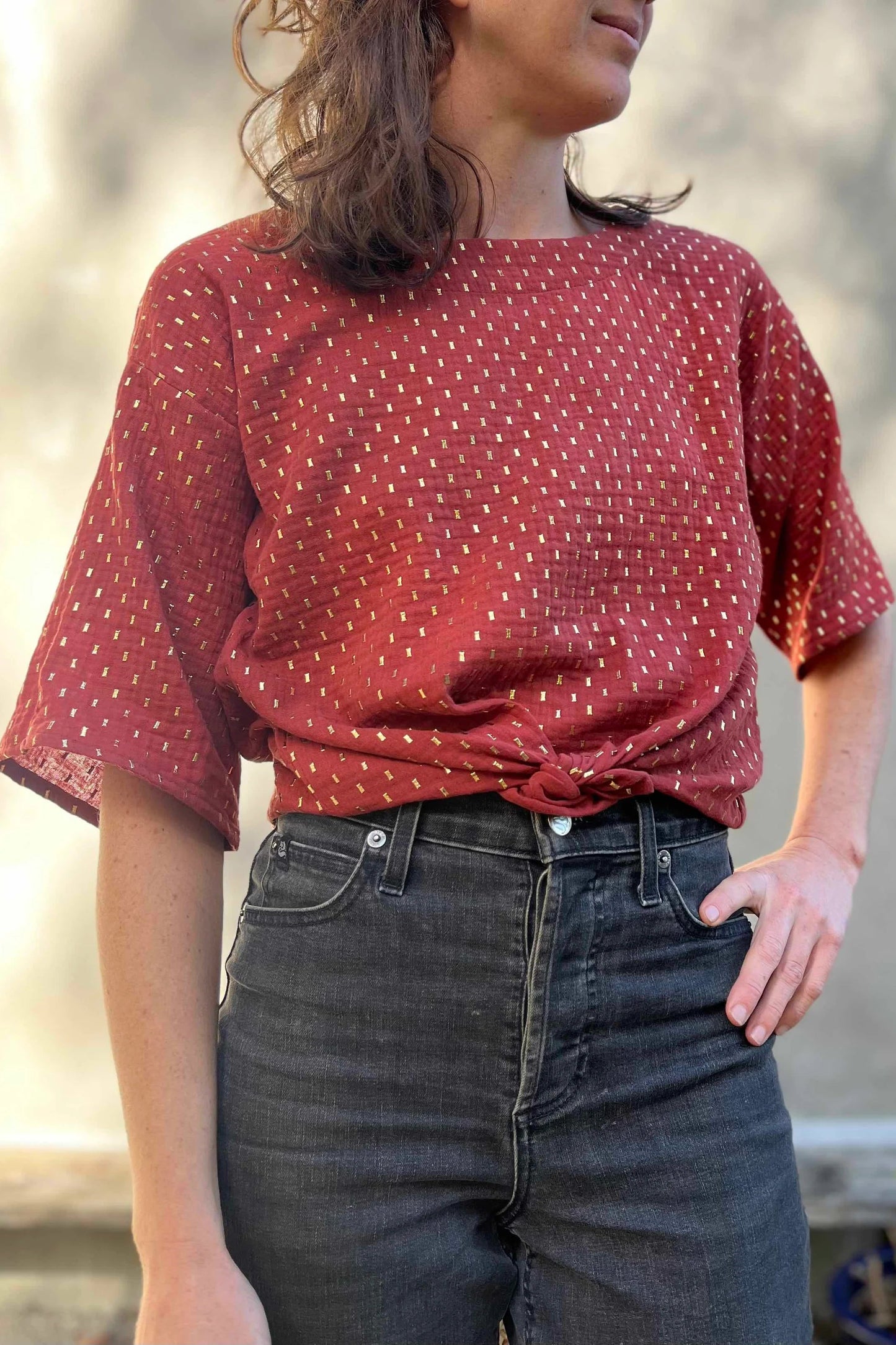 A woman wearing the Abelia Top by Kazak in Golden Tomettes, a boxy top with a wide neck and 3/4 kimono sleeves featuring gold flecks on a red base. She is wearing it with jeans and standing outside.