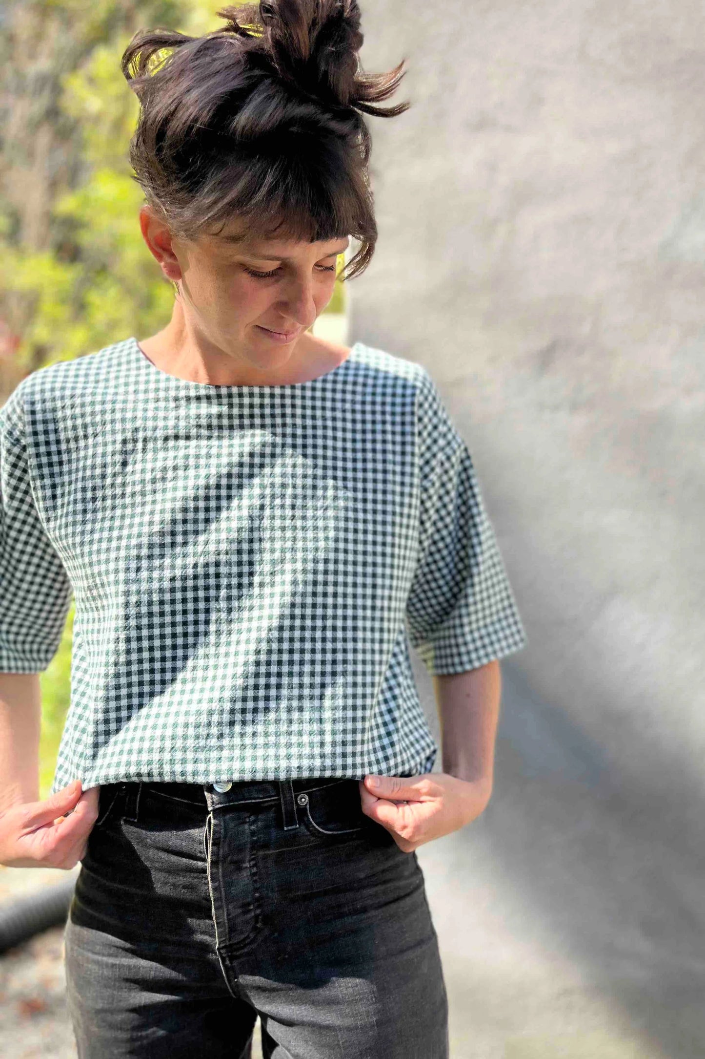 A woman wearing the Abelia Top by Kazak in Forest Vichy, a boxy shirt with a wide neck and 3/4 kimono sleeves. She is wearing it with grey jeans and standing outside against a grey wall.