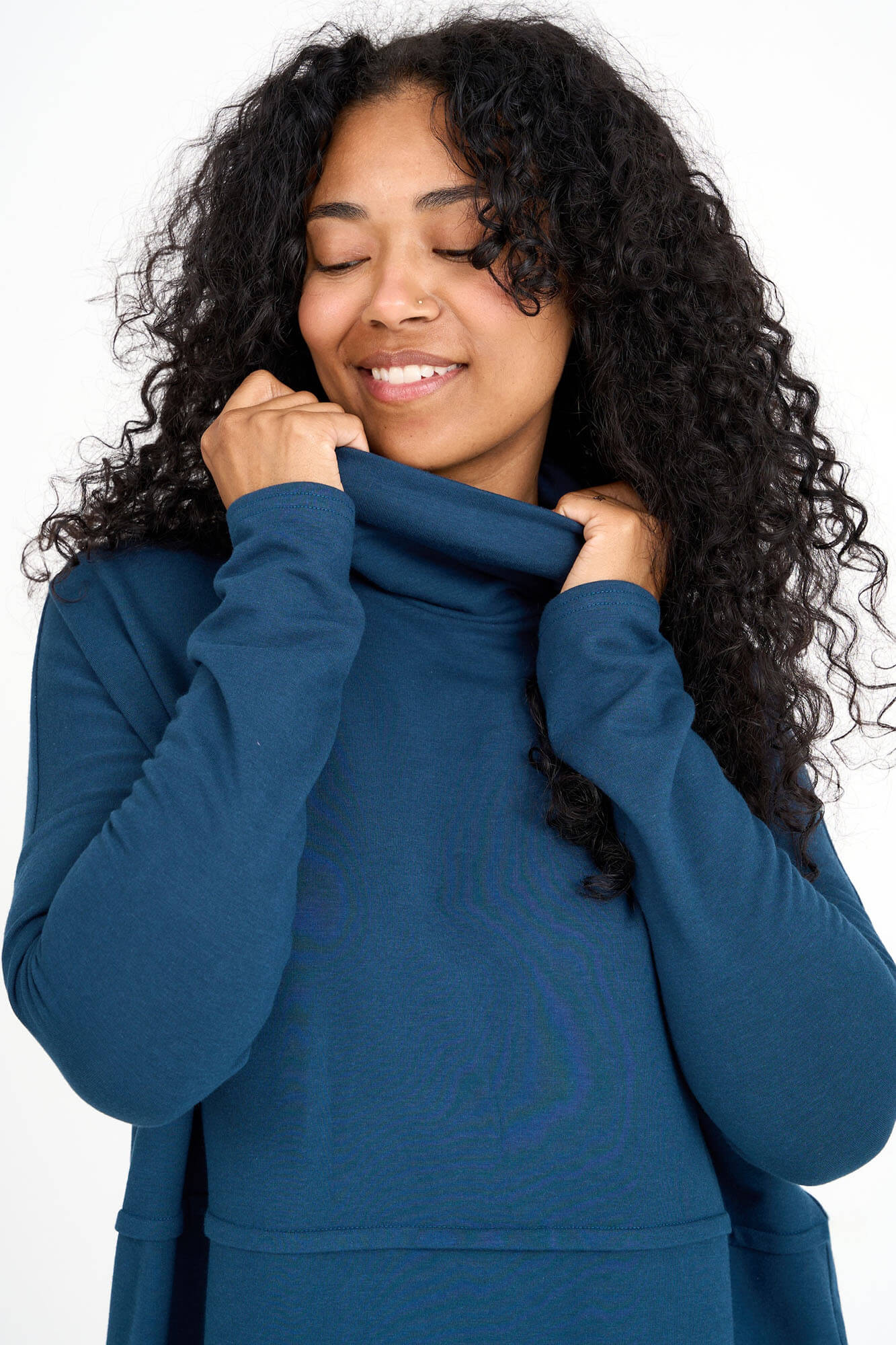Close up of a woman wearing the Abigail Turtleneck by Advika in Bluebell, a loose fitting turtleneck top with dropped sleeves, a seam across the middle, and a rounded hem. She is standing in front of a white background.