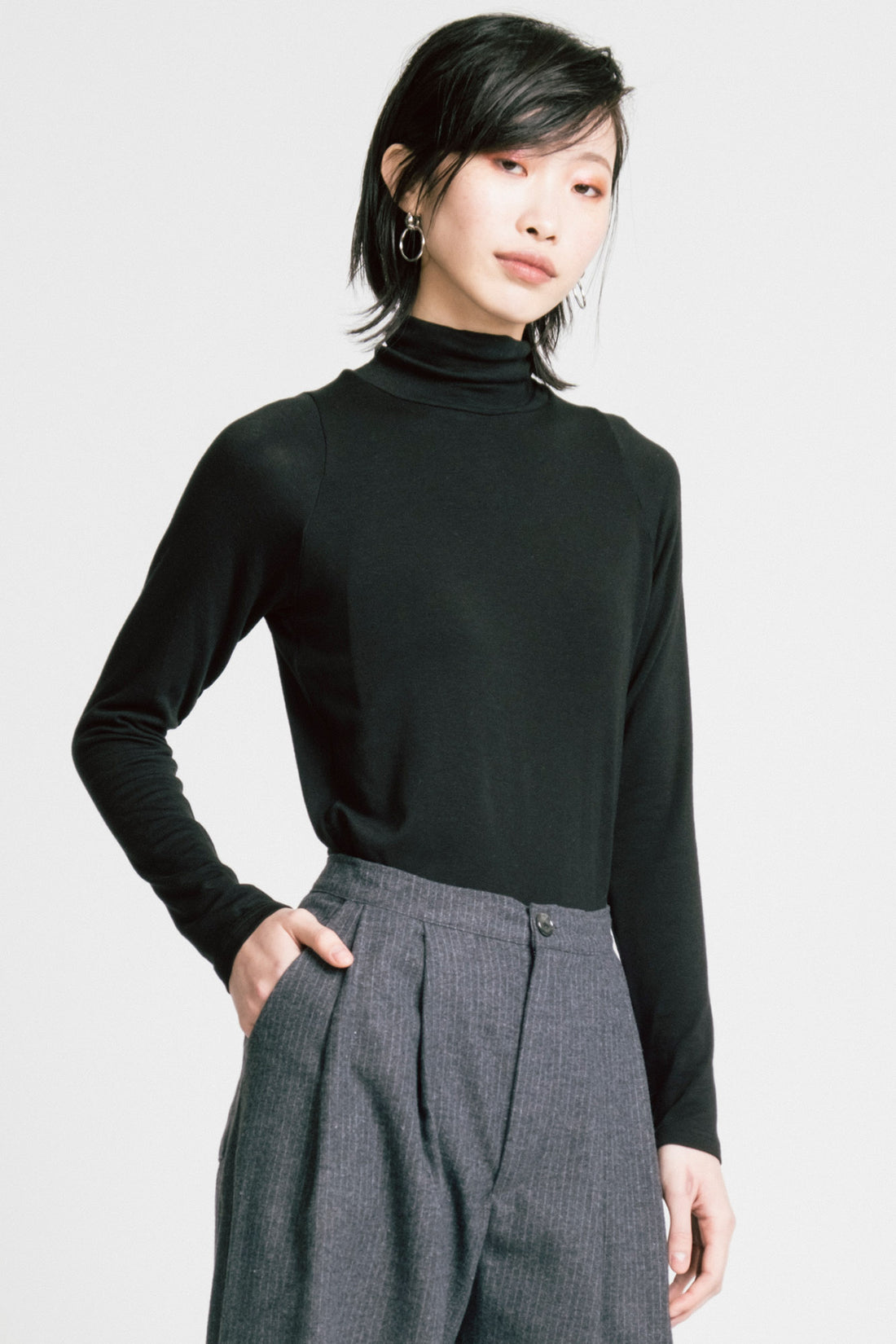 A woman wearing the Aches Turtleneck by Allison Wonderland in Black, a slim fitting classic turtleneck. She is wearing it with the Memory Pants in Denim Stripe and standing in front of a white background.