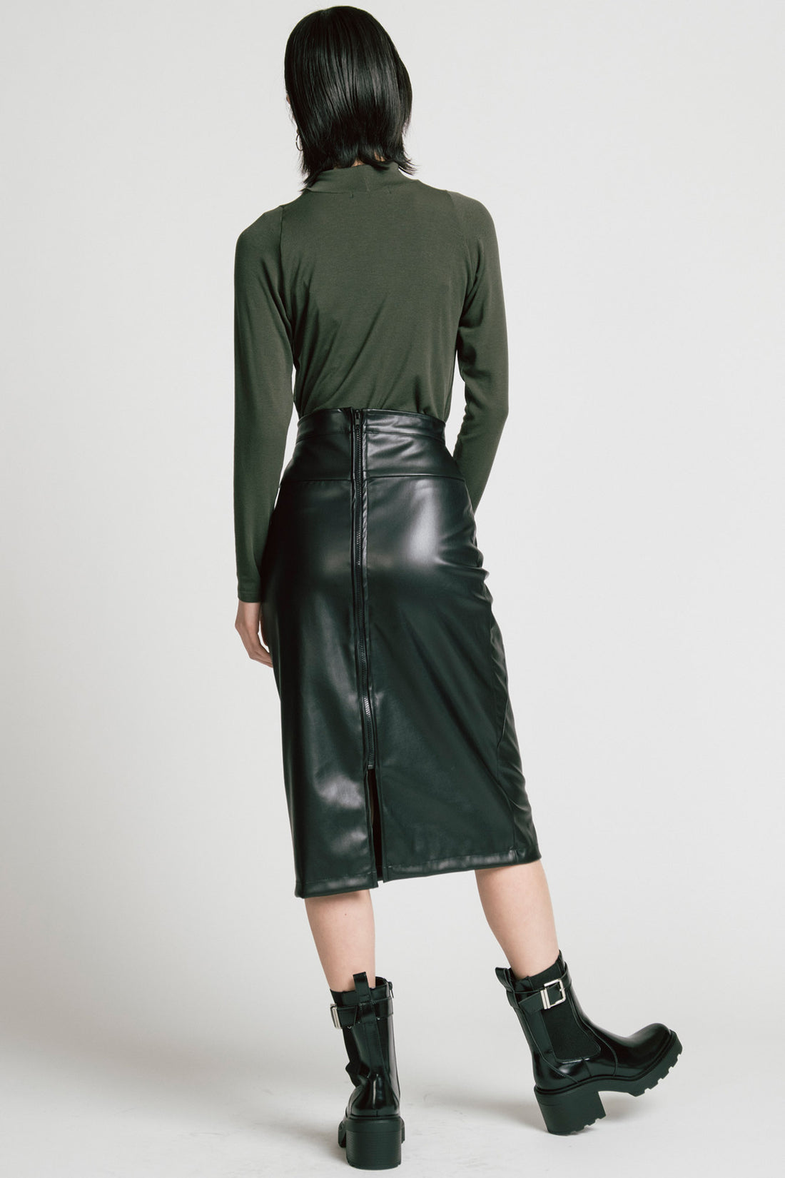 Back view of a woman wearing the Aches Turtleneck by Allison Wonderland in Green, a slim fitting classic turtleneck. She is wearing it with the Desire Skirt in Black and standing in front of a white background.