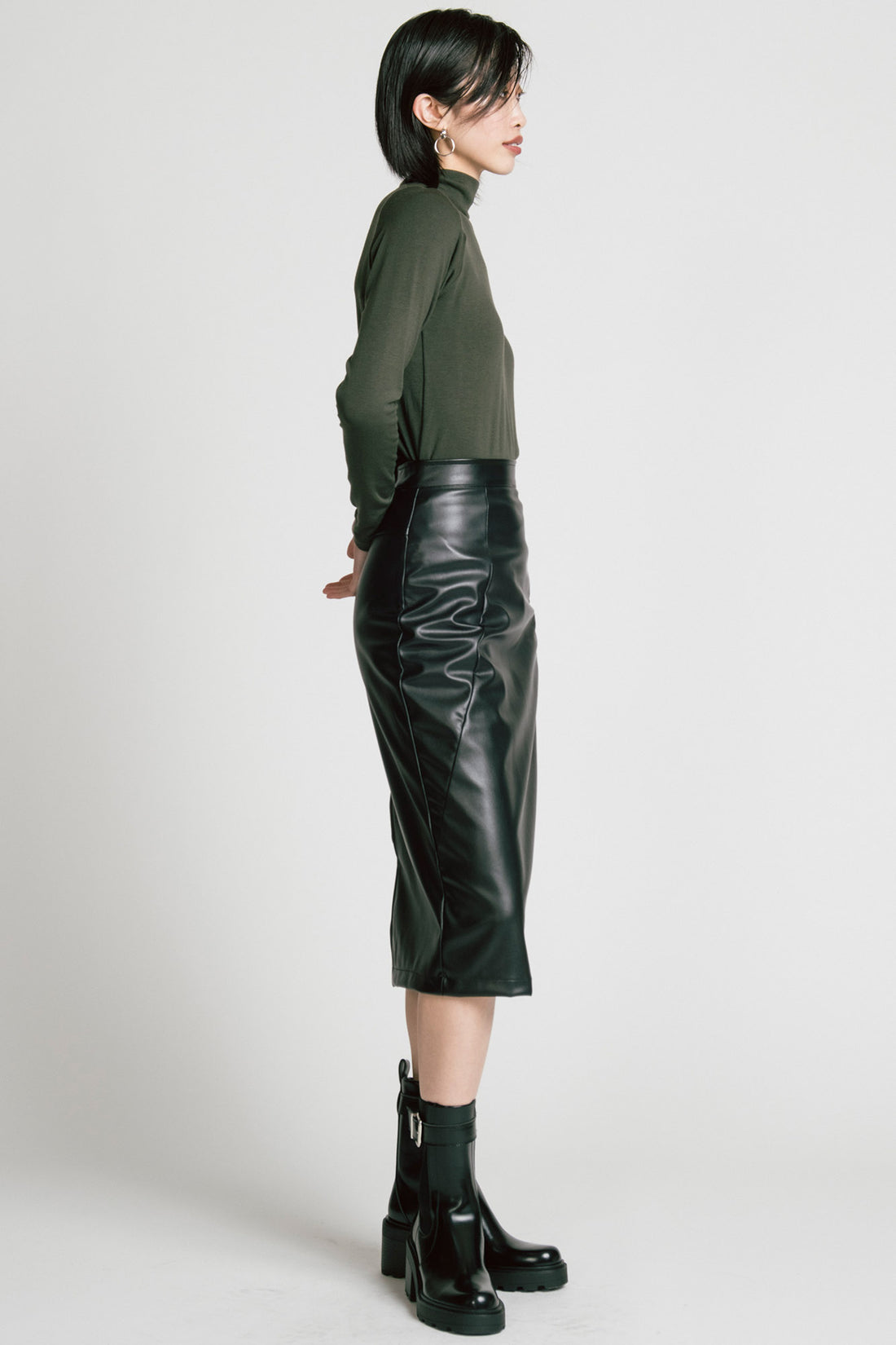 Side view of a woman wearing the Aches Turtleneck by Allison Wonderland in Green, a slim fitting classic turtleneck. She is wearing it with the Desire Skirt in Black and standing in front of a white background.
