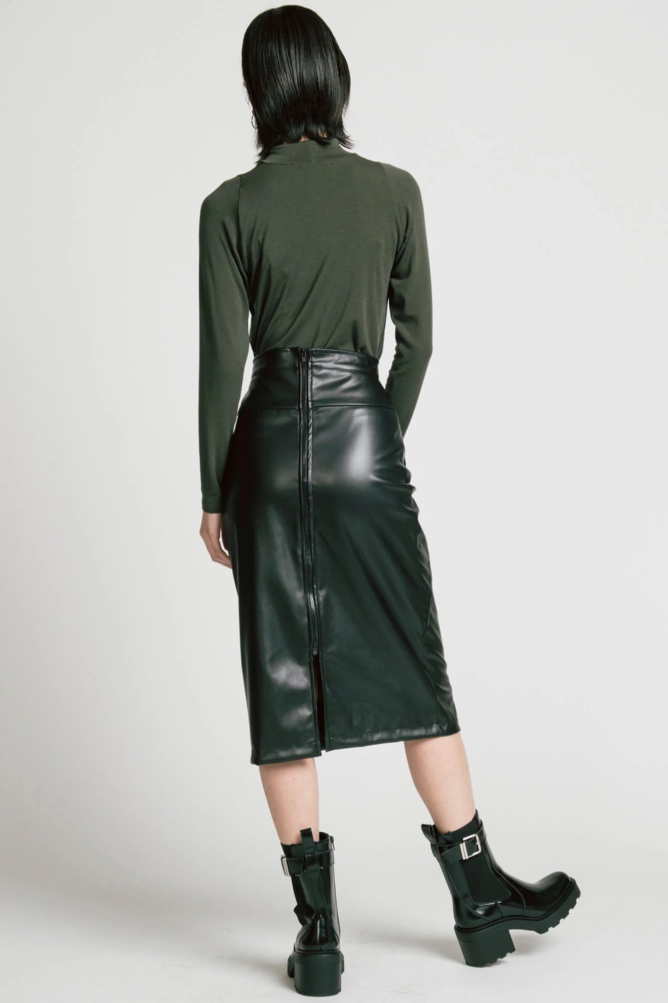 Back view of a woman wearing the Desire Skirt by Allison Wonderland in Black faux leather, a midi-length pencil skirt with a two-way back zipper. She is wearing it with a green turtleneck and standing in front of a white background.