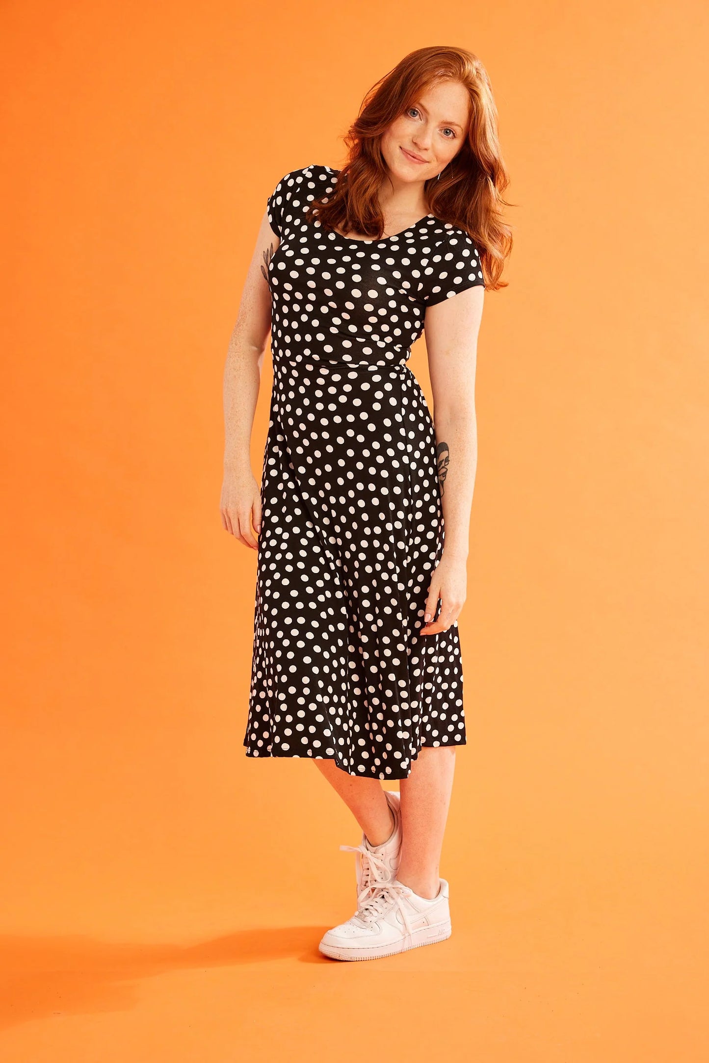 A woman wearing the Rose Dress by Canopée in Black and White Polka Dots, with short sleeves, a round neck, a cross-over back, and a fitted bodice with a below-the-knee skirt. She is standing in front of an orange background.