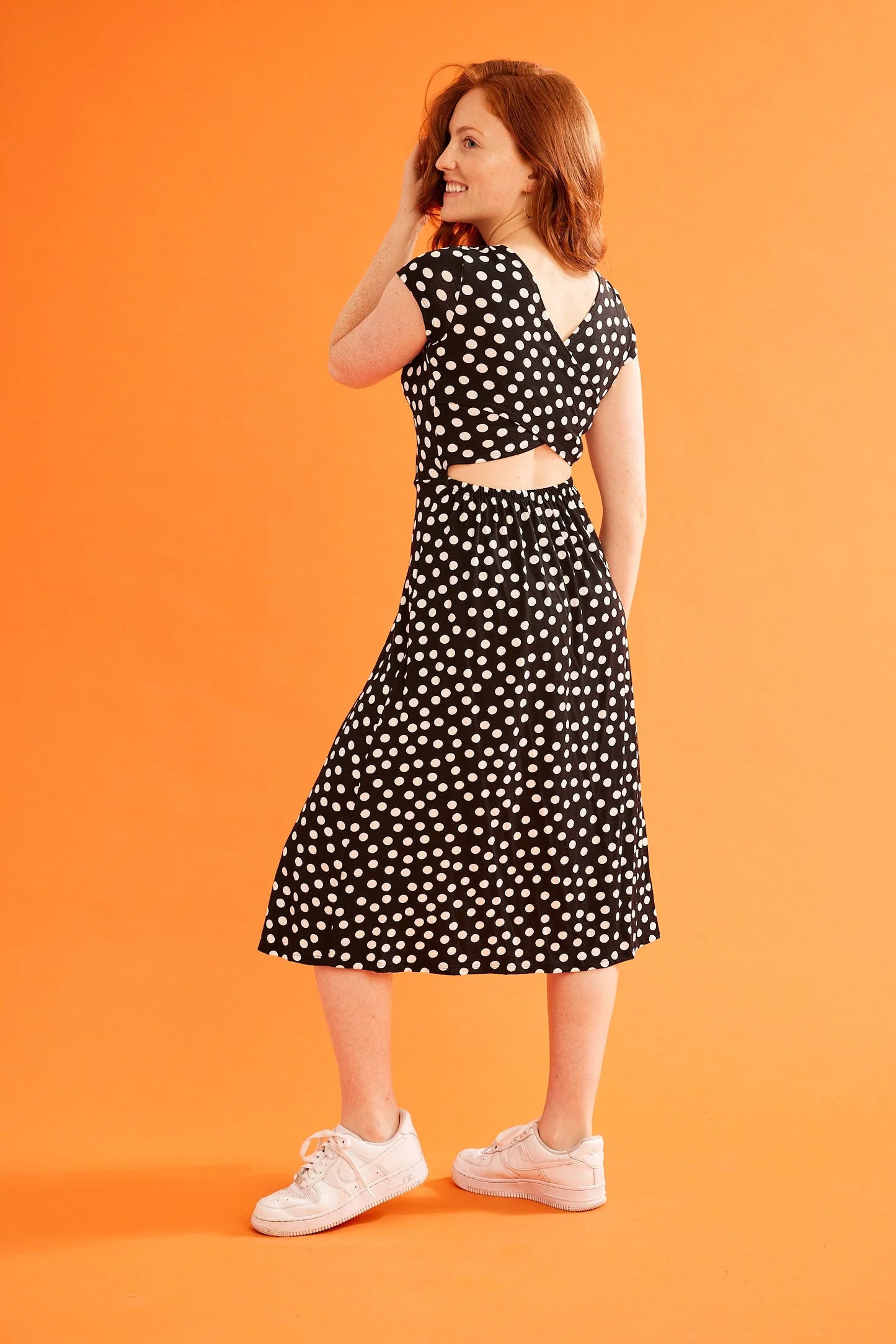 Back view of a woman wearing the Rose Dress by Canopée in Black and White Polka Dots, with short sleeves, a round neck, a cross-over back, and a fitted bodice with a below-the-knee skirt. She is standing in front of an orange background.