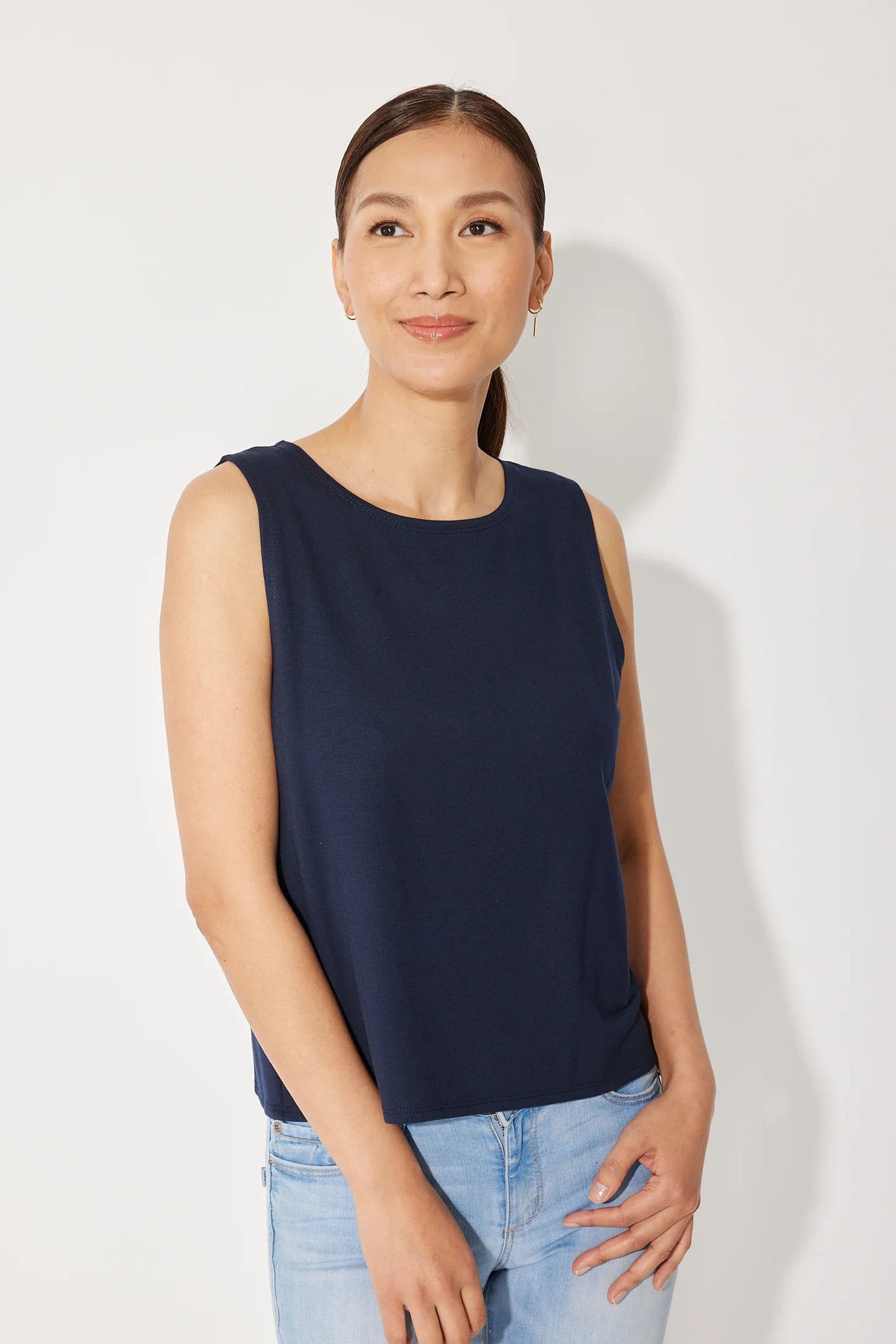 A woman wearing the Baie Cami by Canopée in Navy, a bamboo French Terry tank with a high neck and loose fit. She is wearing it with jeans and standing in front of a white background.