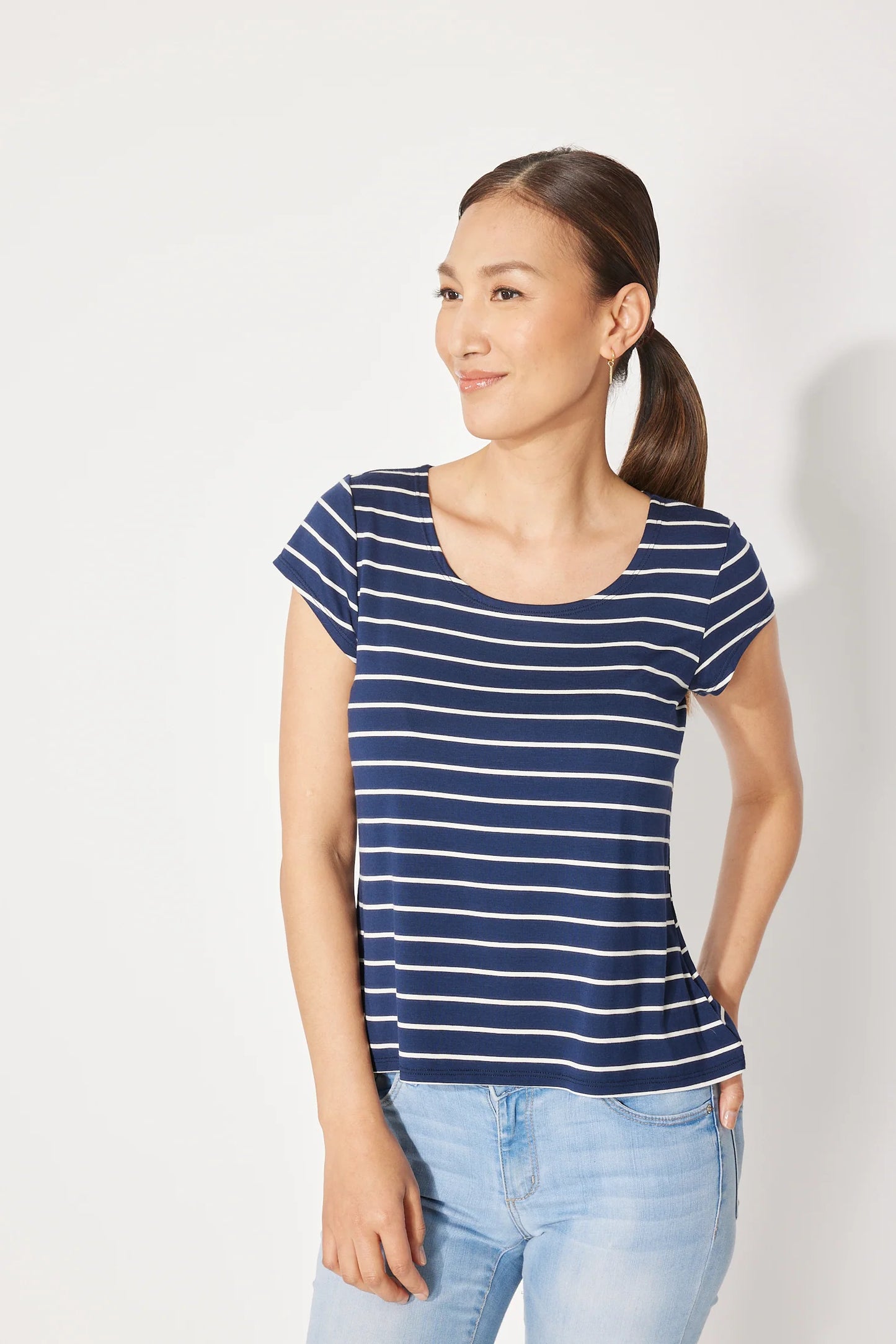A woman wearing the Fjord T-shirt by Canopée in Navy/White stripe, a short-sleeved tee with a wide scooped neck and a slightly loose fit. She is wearing it with jeans and standing in front of a white background.