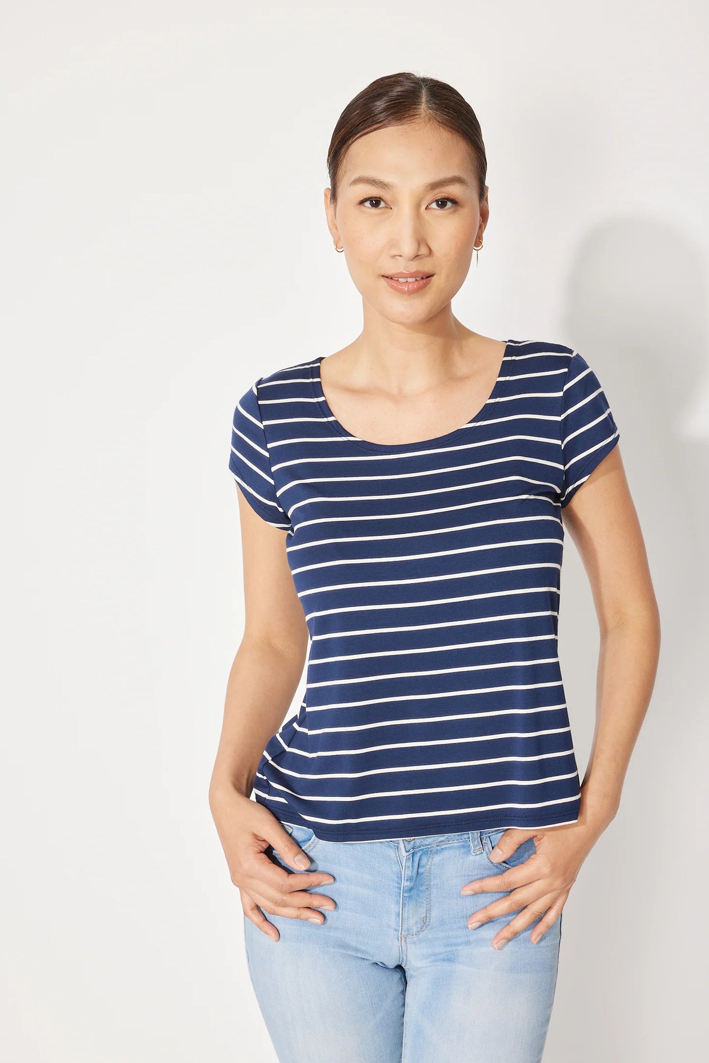 A woman wearing the Fjord T-shirt by Canopée in Navy/White stripe, a short-sleeved tee with a wide scooped neck and a slightly loose fit. She is wearing it with jeans and standing in front of a white background.