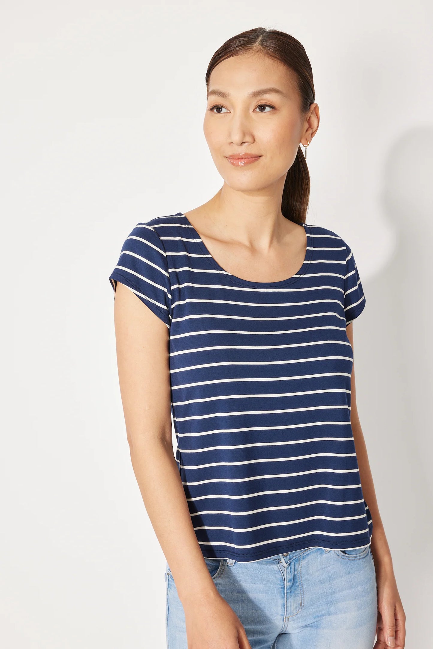 A woman wearing the Fjord T-shirt by Canopée in Navy/White stripe, a short-sleeved tee with a wide scooped neck and a slightly loose fit. She is wearing it with jeans and standing in front of a white background.