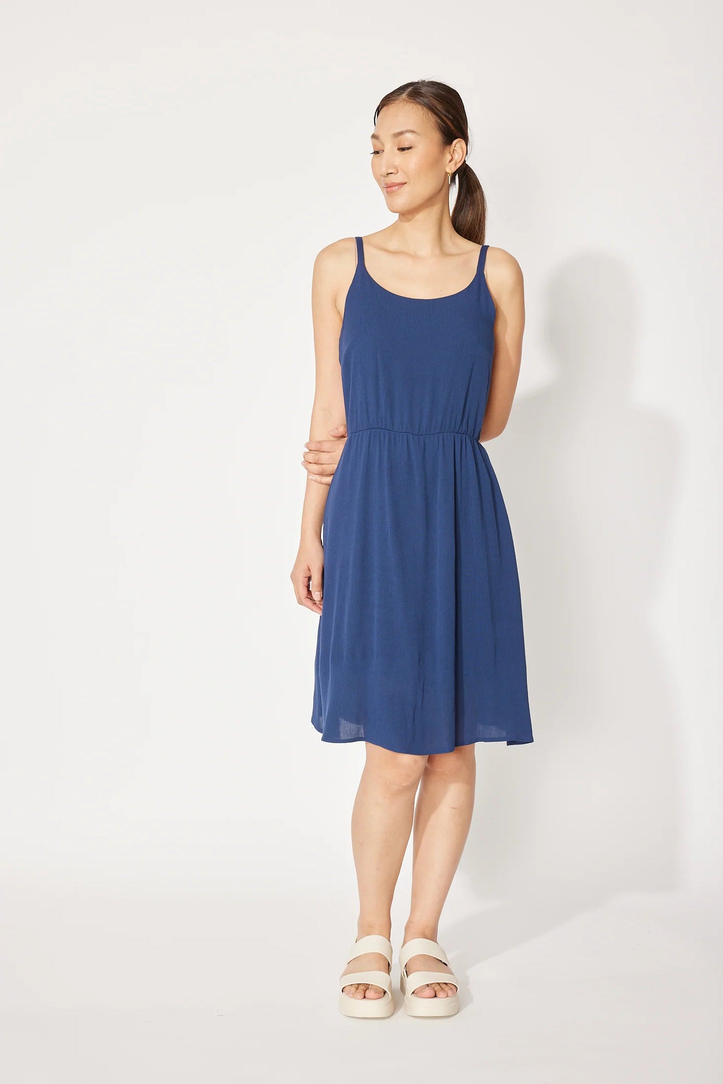 A woman wearing the Wildflower Dress by Cherry Bobin in Navy, a light voile dress with thin straps, an elastic waist, and a full knee-length skirt. She is standing in front of a white background.