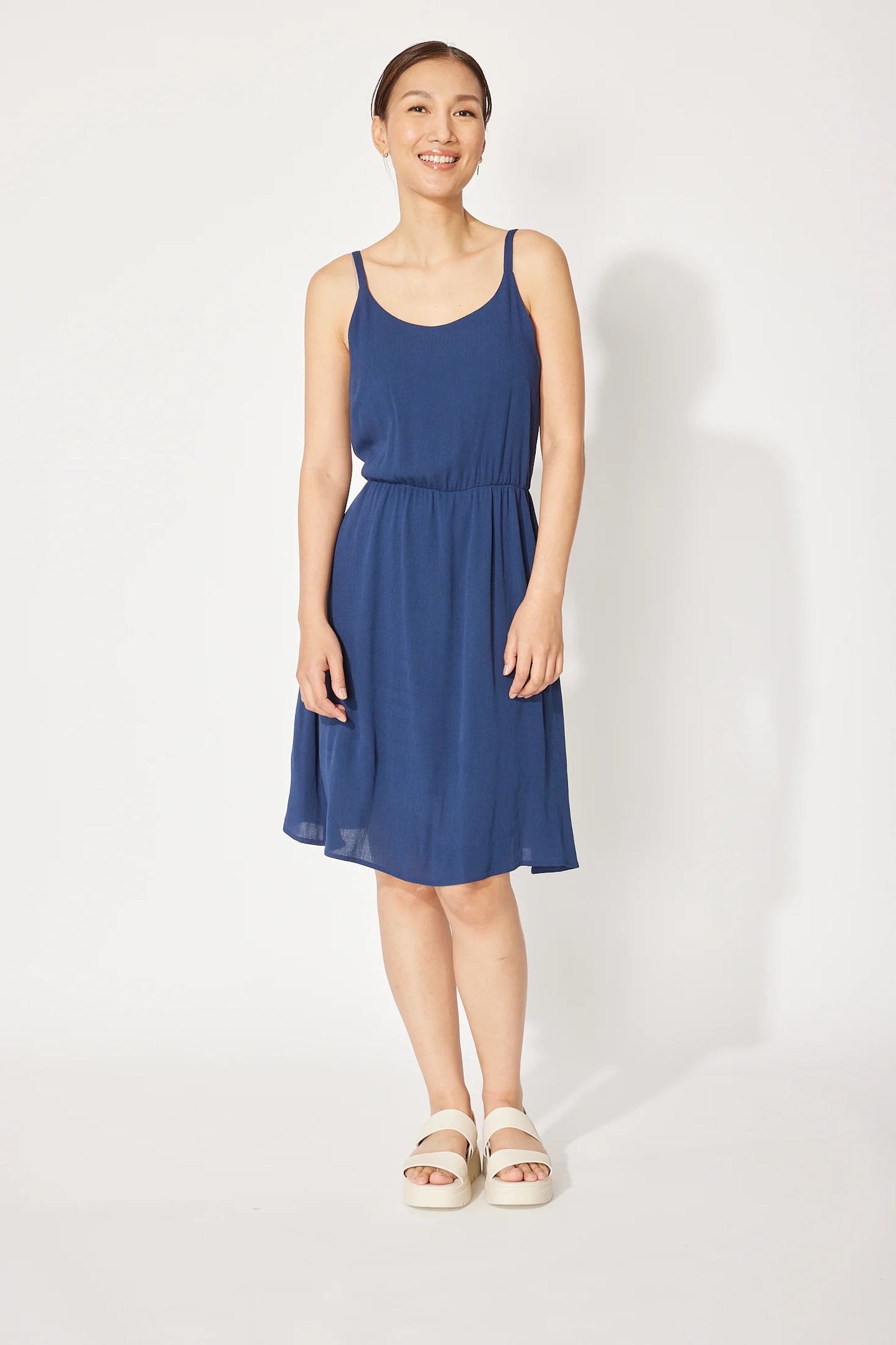 A woman wearing the Wildflower Dress by Cherry Bobin in Navy, a light voile dress with thin straps, an elastic waist, and a full knee-length skirt. She is standing in front of a white background.