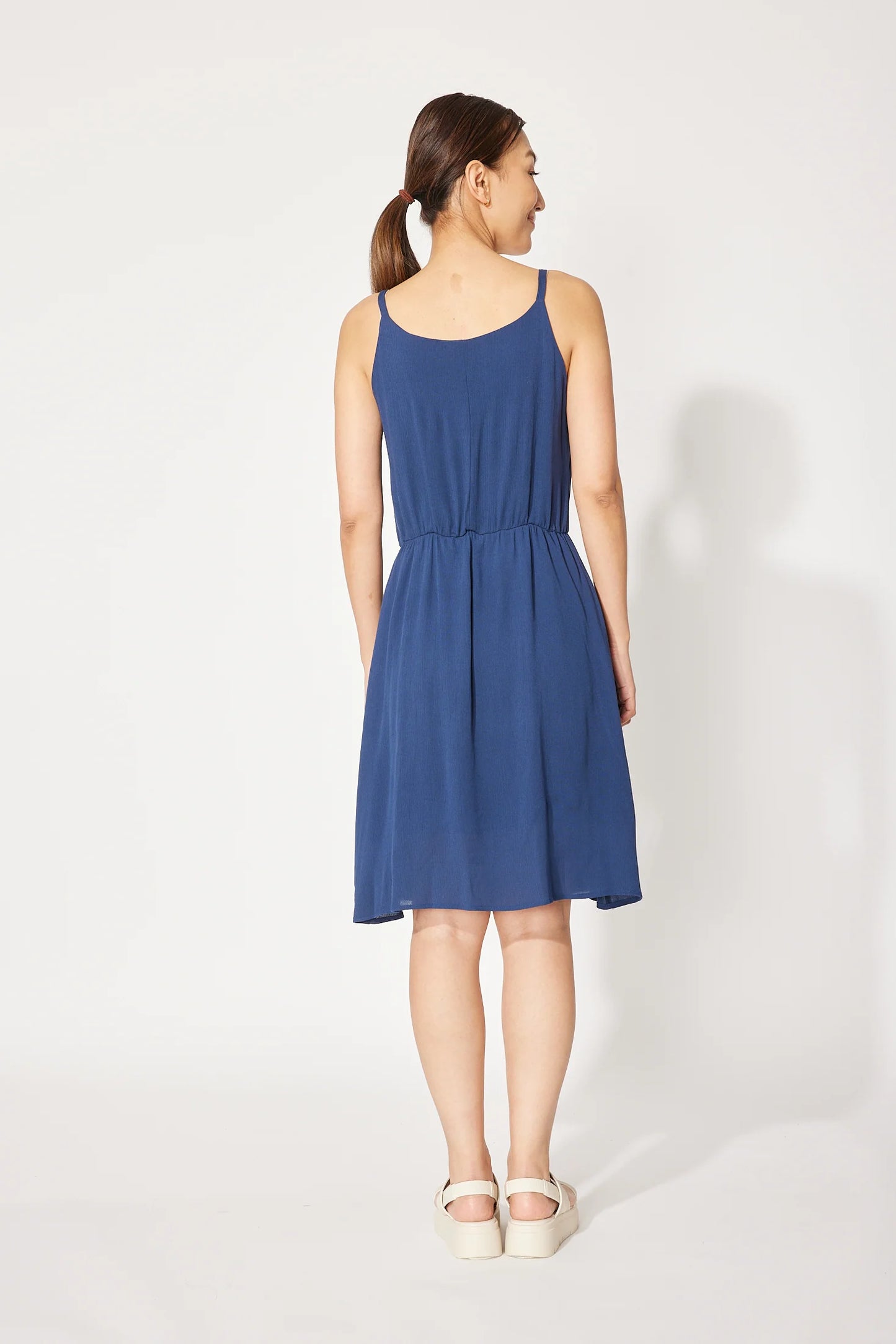Back view of a woman wearing the Wildflower Dress by Cherry Bobin in Navy, a light voile dress with thin straps, an elastic waist, and a full knee-length skirt. She is standing in front of a white background.