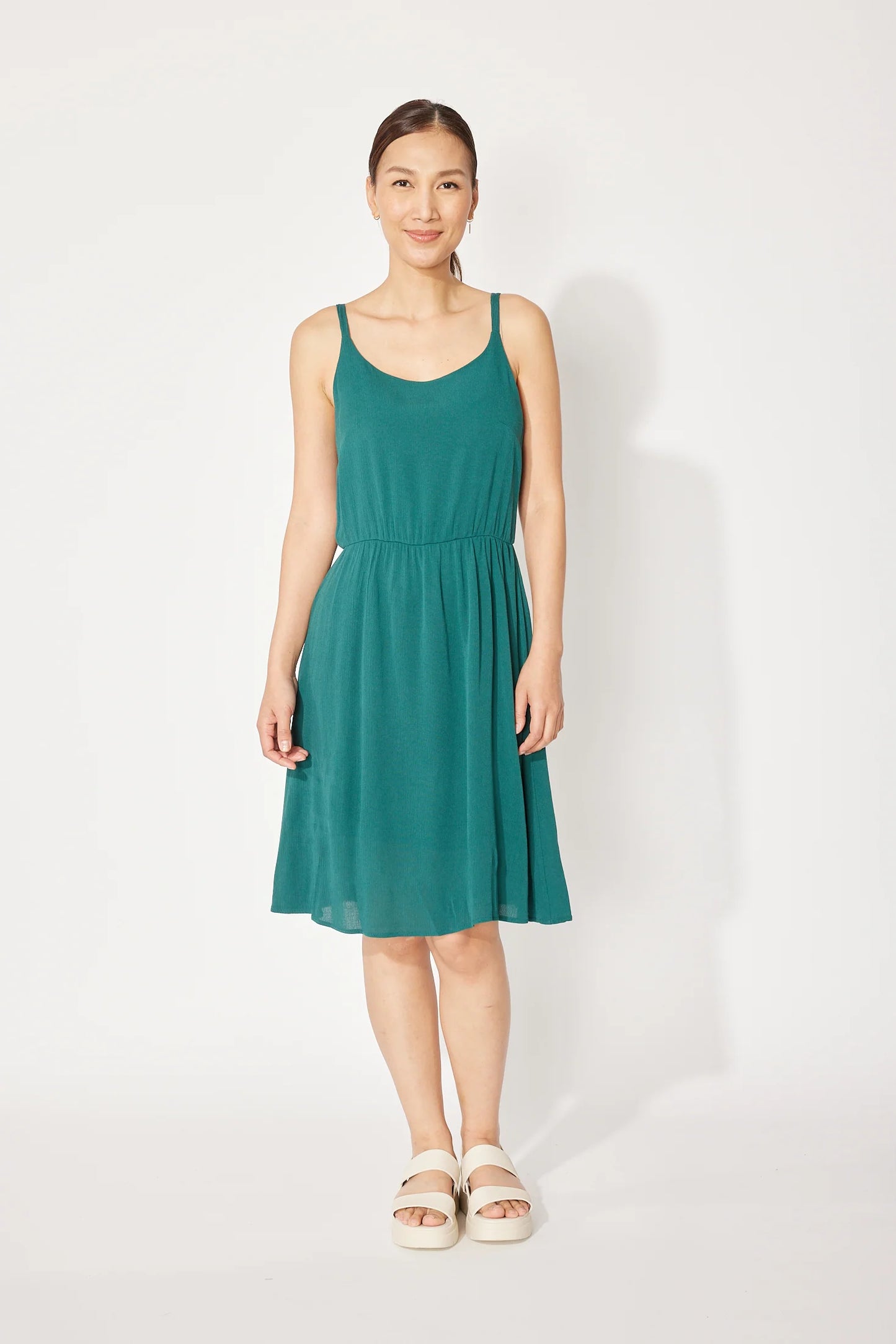 A woman wearing the Wildflower Dress by Cherry Bobin in Turquoise, a light voile dress with thin straps, an elastic waist, and a full knee-length skirt. She is standing in front of a white background.