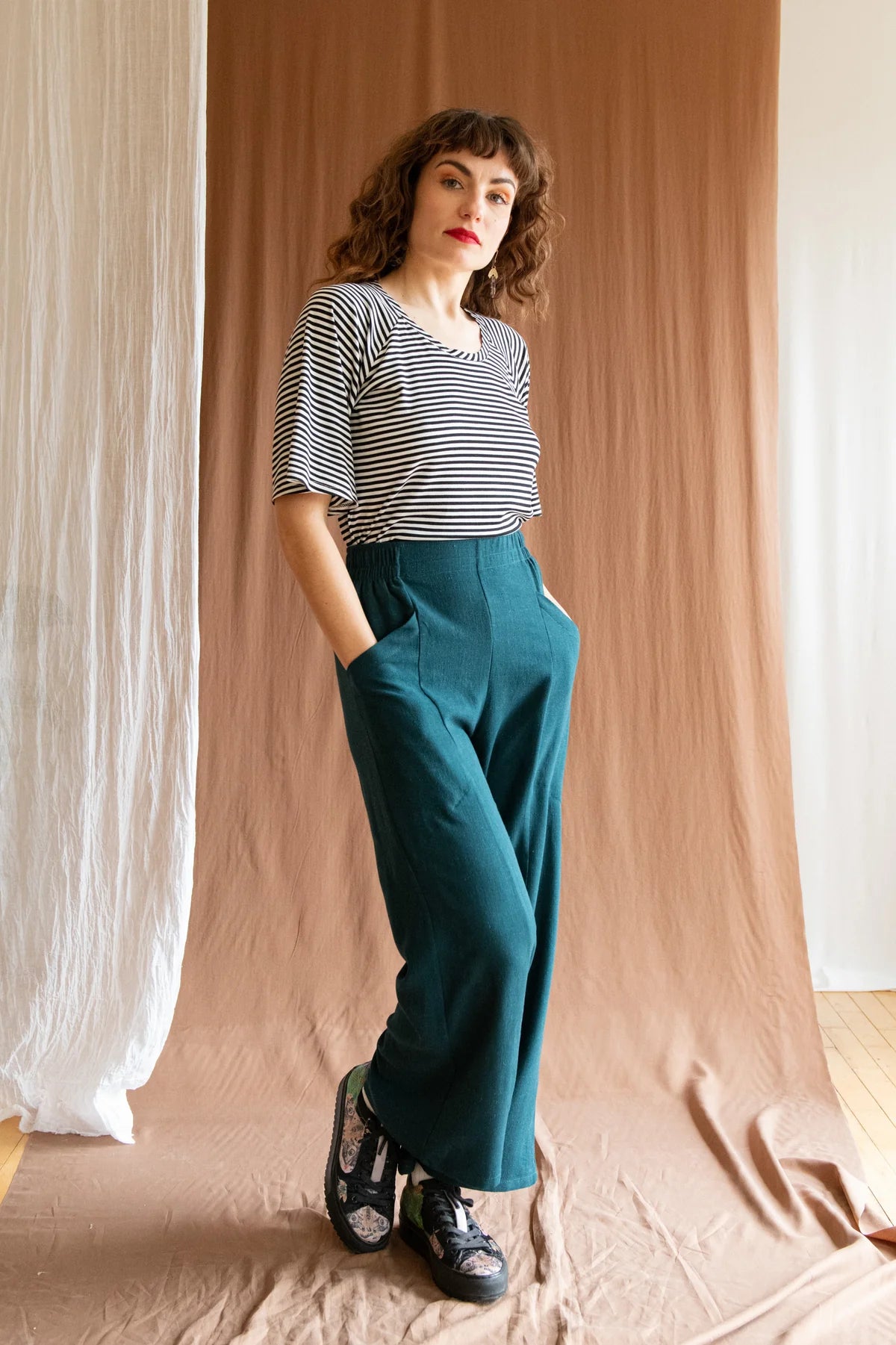 A woman wearing the Montebelluna Top by Kazak in Navy Stripe, featuring raglan style, flared elbow-length sleeves, and a scooped neck. She is wearing it with the Padua Pants in Forest, and is standing in front of a brown curtain.