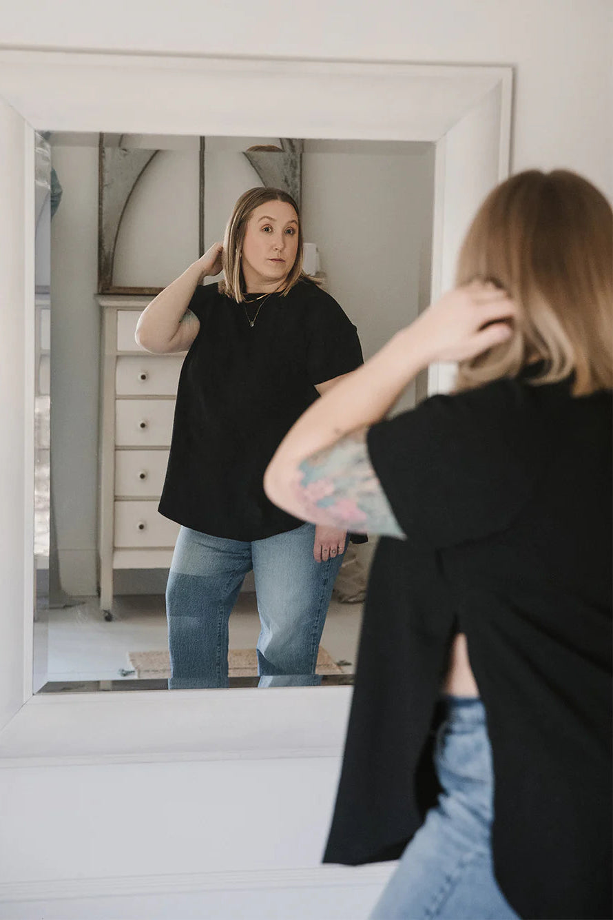 A woman wearing the East End Tee by Blondie in Black and looking at her reflection in a mirror. The top has a round neck, short raglan sleeves, and long slits up the sides.