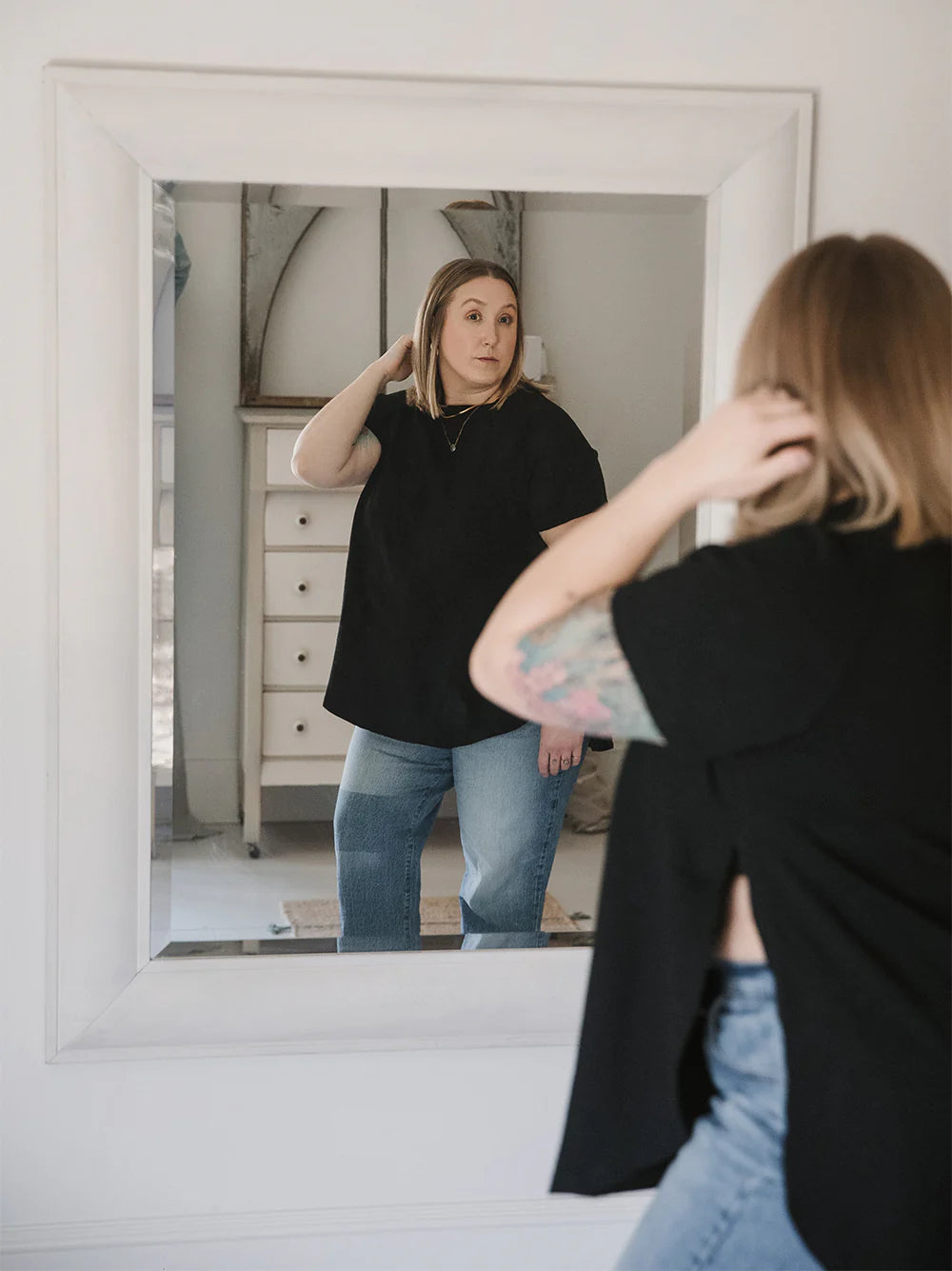 A woman wearing the East End Tee by Blondie in Black and looking at her reflection in a mirror. The top has a round neck, short raglan sleeves, and long slits up the sides.