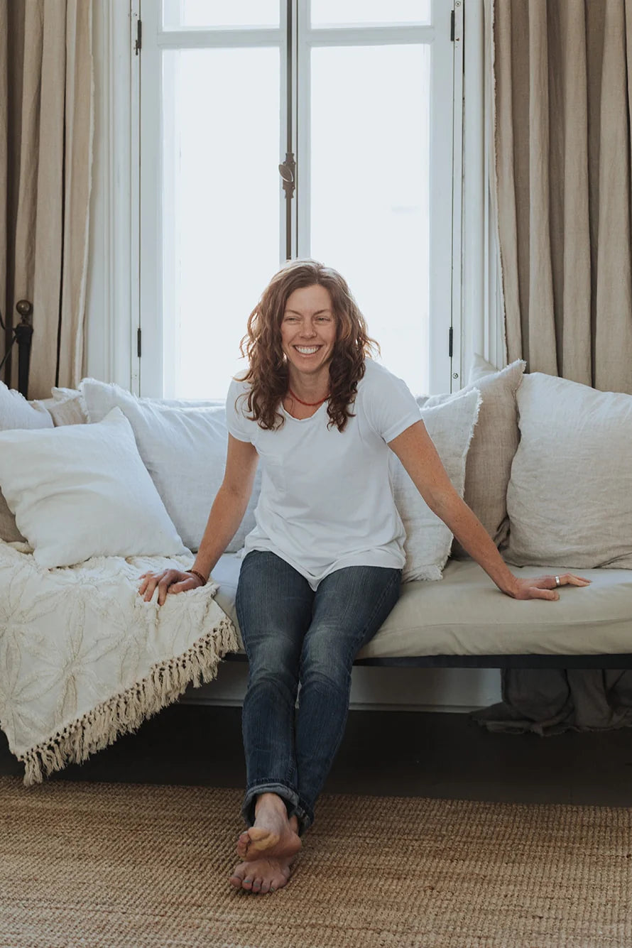 A woman wearing the Janis Tee by Blondie in White, with a round neck, short sleeves, breast pocket, and hi-low hemline. She is wearing it with jeans skirt and sitting on a couch.