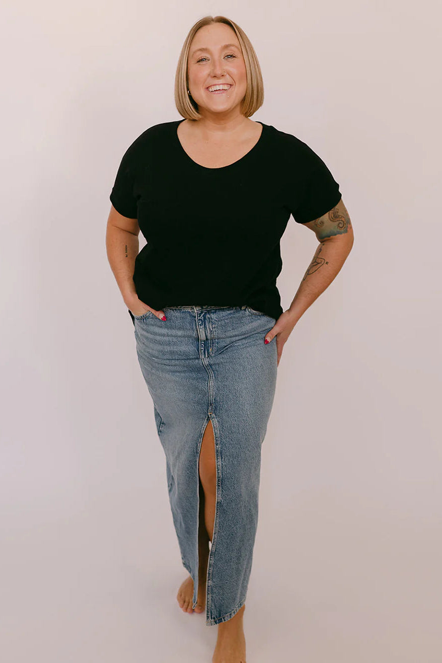 A woman wearing the Janis Tee by Blondie in Black Slub, with a round neck, short sleeves, breast pocket, and hi-low hemline. She is wearing it with a jean skirt and standing in front of a white background.