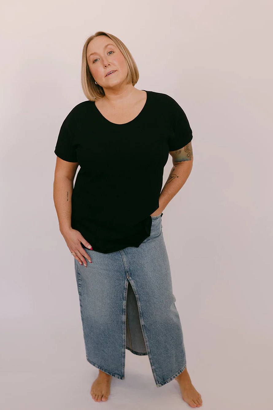 A woman wearing the Janis Tee by Blondie in Black Slub, with a round neck, short sleeves, breast pocket, and hi-low hemline. She is wearing it with a jean skirt and standing in front of a white background.
