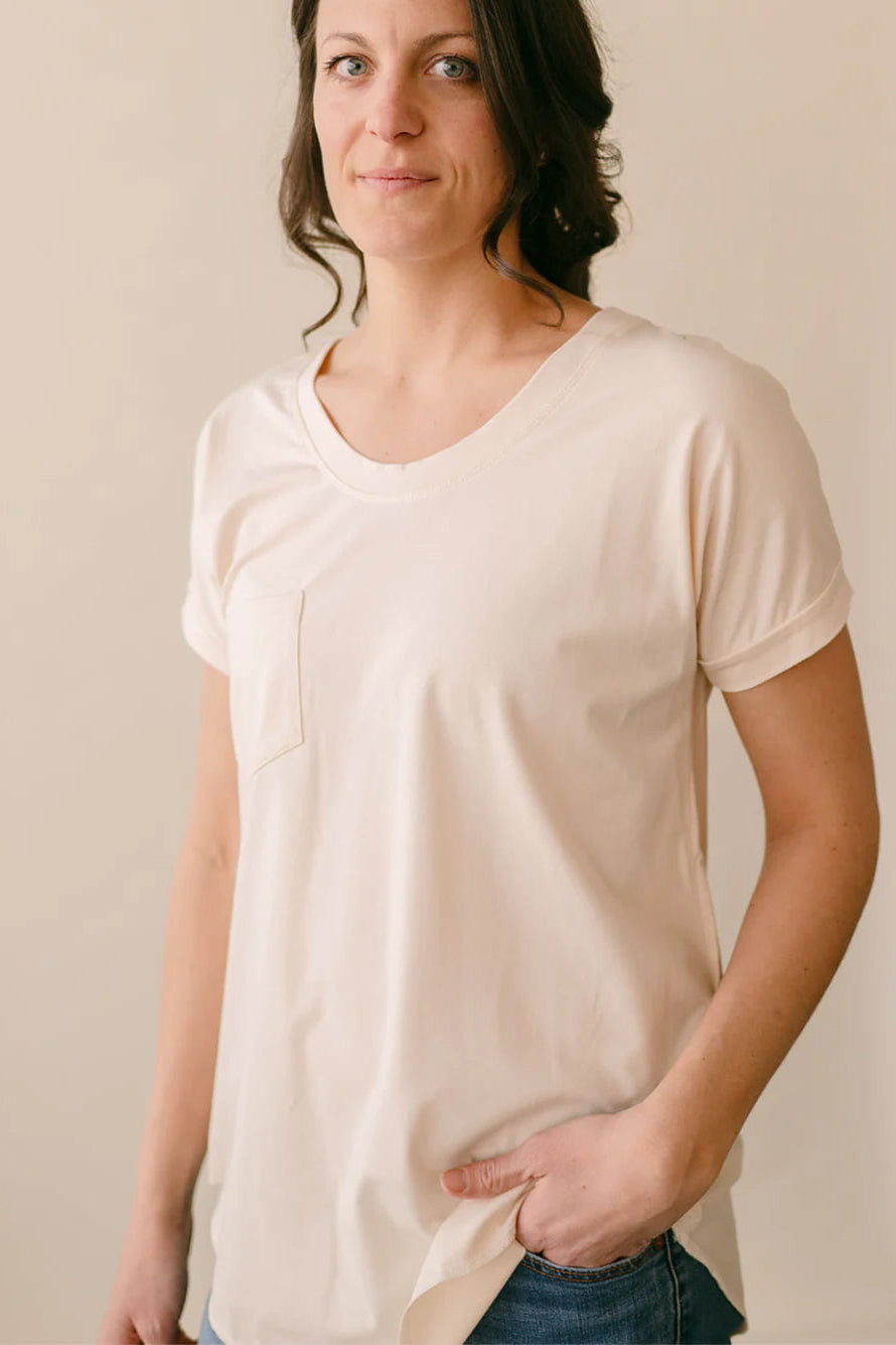 A woman wearing the Janis Tee by Blondie in Ivory, with a round neck, short sleeves, breast pocket, and hi-low hemline. She is wearing it with a jeans. and standing in front of a white background.
