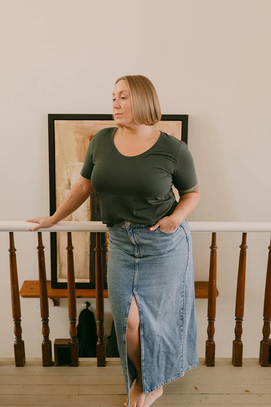 A woman wearing the Rideau Shoreline Tee by Blondie in Pine, with a scoop neck, short sleeves, and a slightly longer length. She is wearing it with a jean skirt and standing in front of a railing.