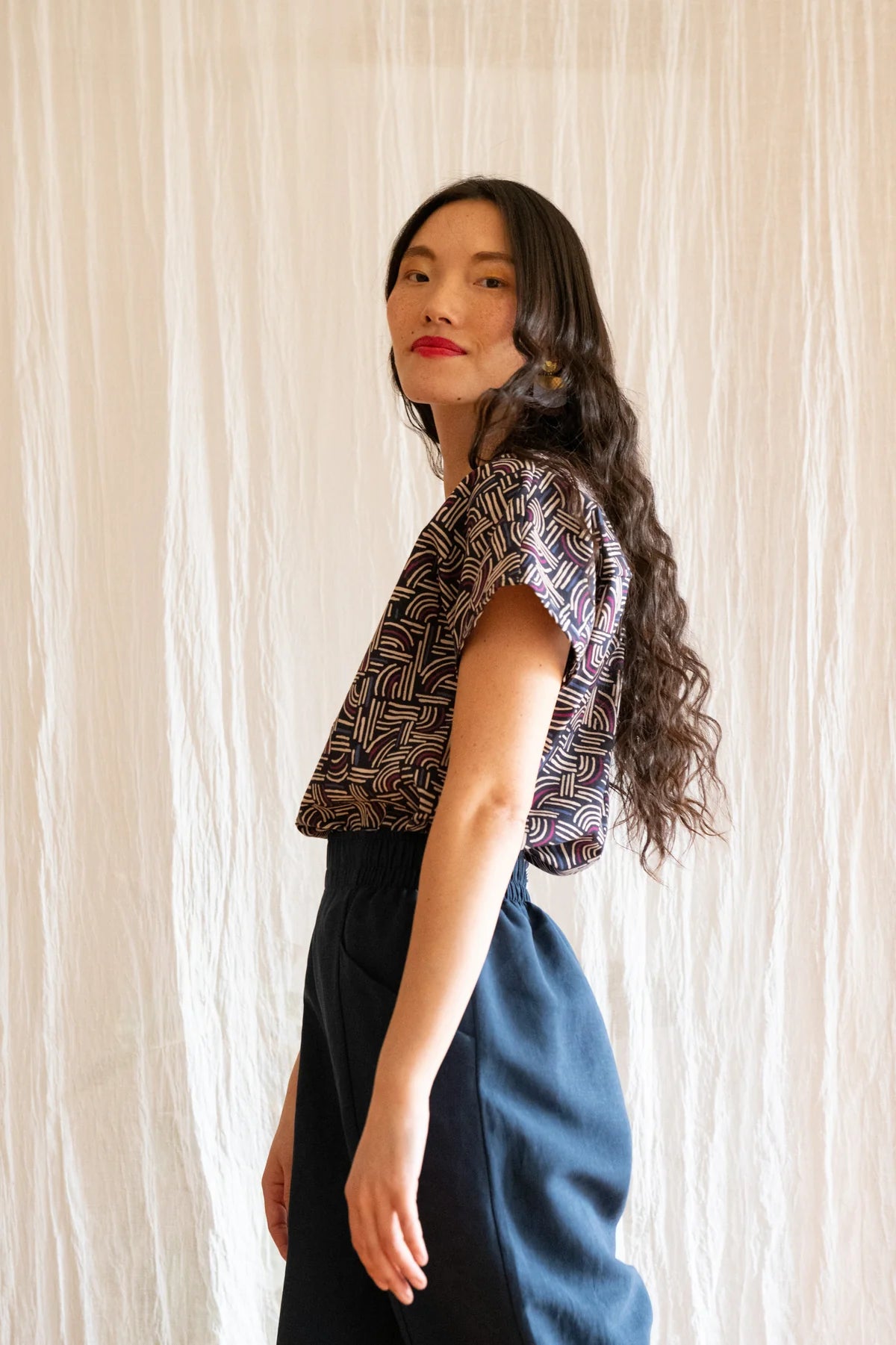 Side 3/4 view of a woman wearing the Padua Pants by Kazak in Indigo, with a high waist, flat waistband at the front and elastic at the bag, wide legs, and large slanted pockets. She is wearing them with the Lucca Top in Night Basketwork and standing in front of a white curtain.