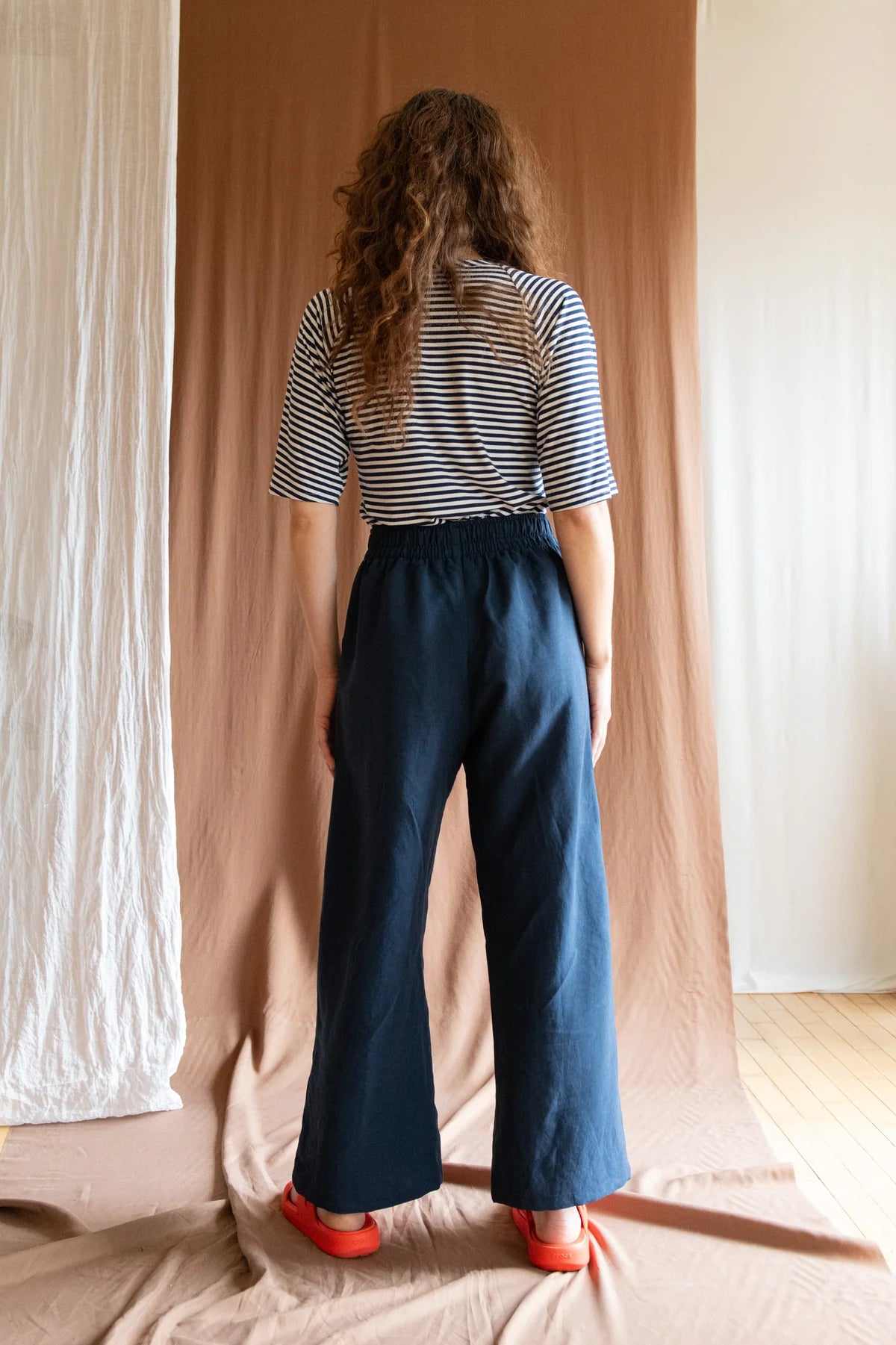 Back view of a woman wearing the Padua Pants by Kazak in Indigo, with a high waist, flat waistband at the front and elastic at the bag, wide legs, and large slanted pockets. She is wearing them with the striped Montebelluna top and standing in front of a brown curtain.