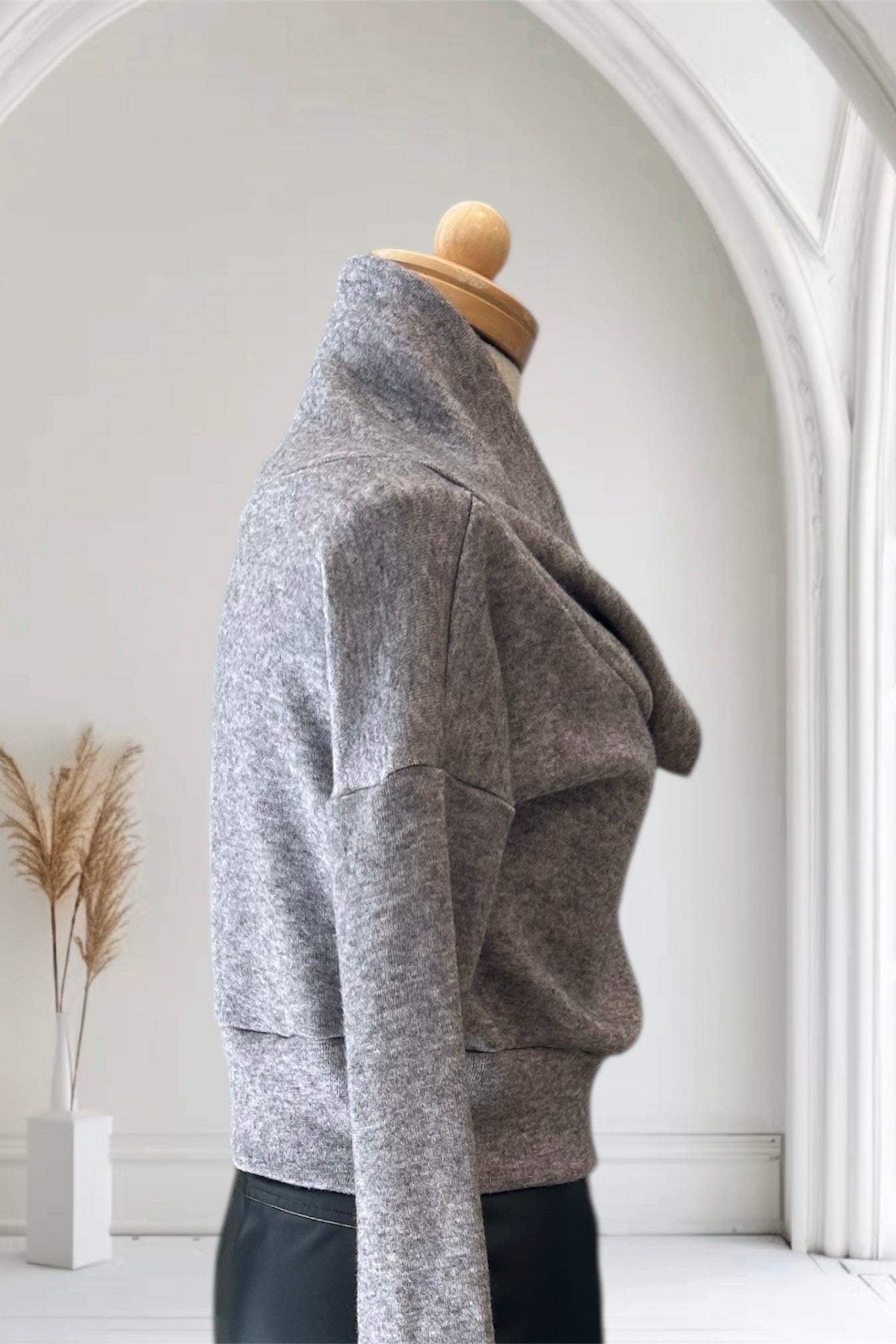 A side view of the Changes Sweater by Allison Wonderland in Light Grey, featuring a large cowl neck, long dropped sleeves, and a wide waistband, is shown with the Desire Skirt in Black against a white arched backdrop.