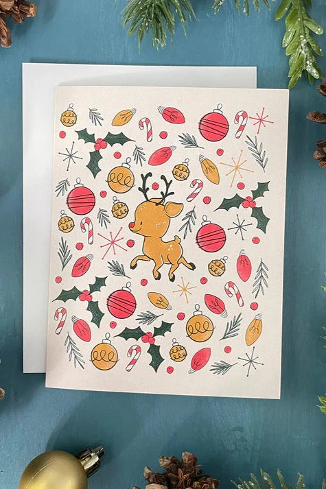 A card from a box set by Carabara Designs, featuring a Midcentury Reindeer pattern on a white background, is shown against a teal background with holiday decor around it.