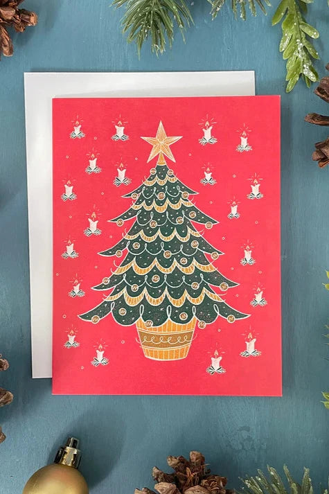 A card from a box set by Carabara Designs, featuring a Midcentury Christmas Tree design on a red background, is shown against a teal background with holiday decor around it.